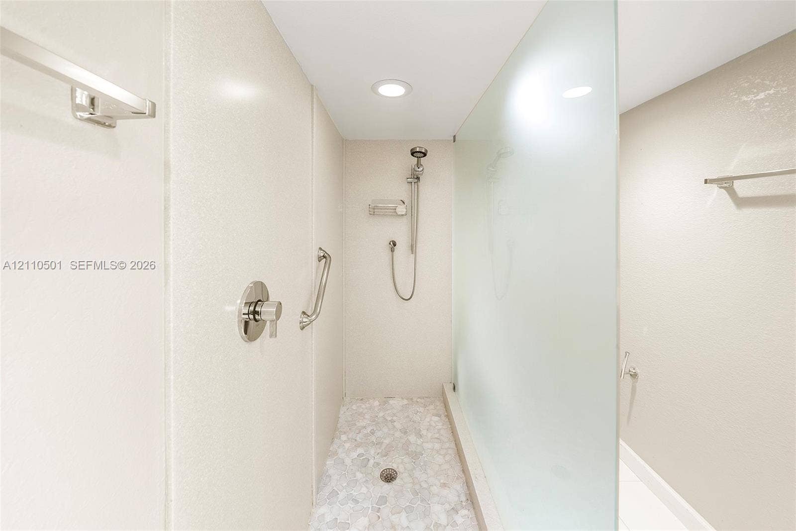 Walk-in shower featuring a frosted glass panel, pebble-stone flooring, brushed-nickel handheld shower head, and wall-mounted metal support bars