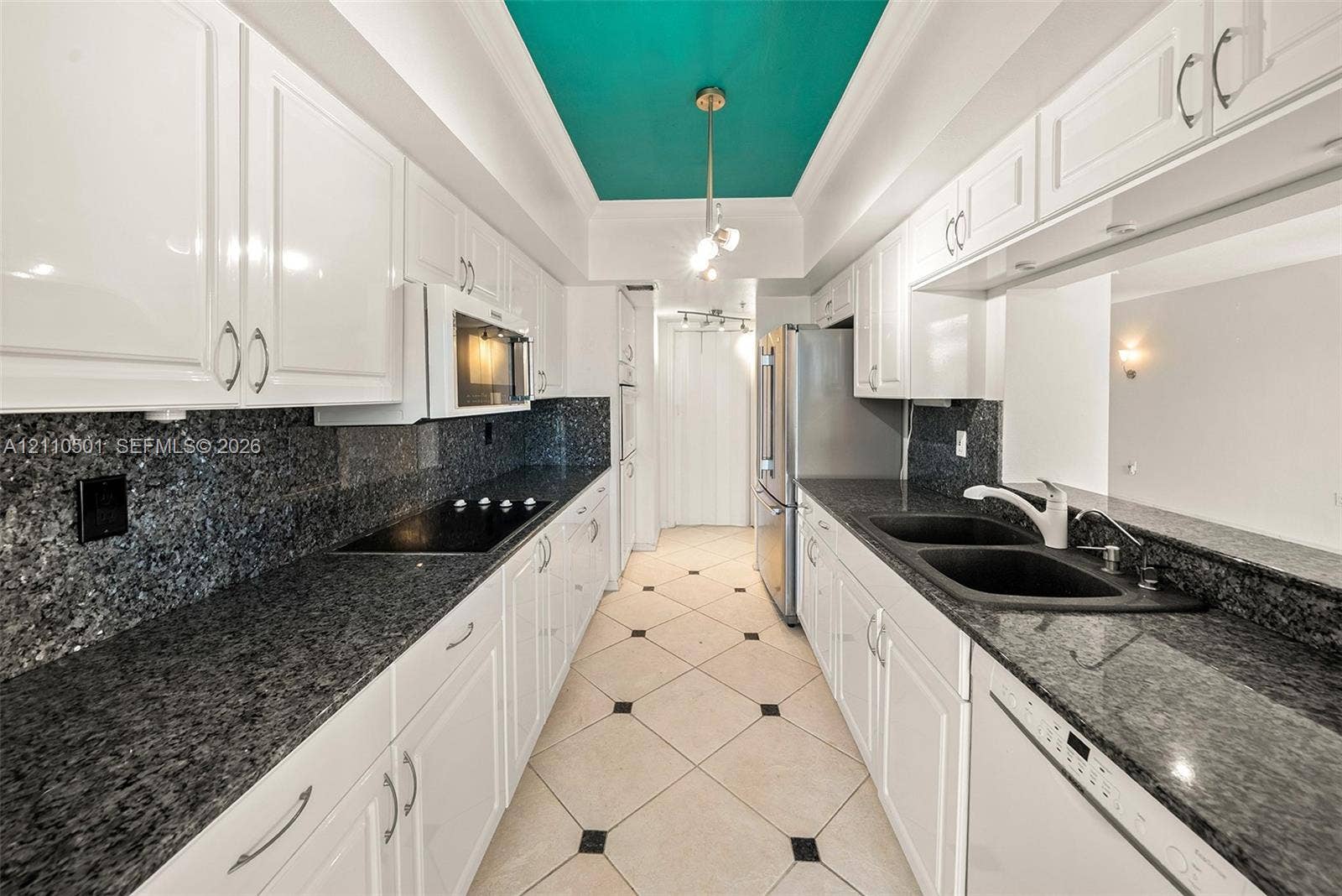 Galley kitchen featuring white cabinetry, dark stone countertops, tile flooring with inset accents, stainless steel appliances, and a recessed tray ceiling