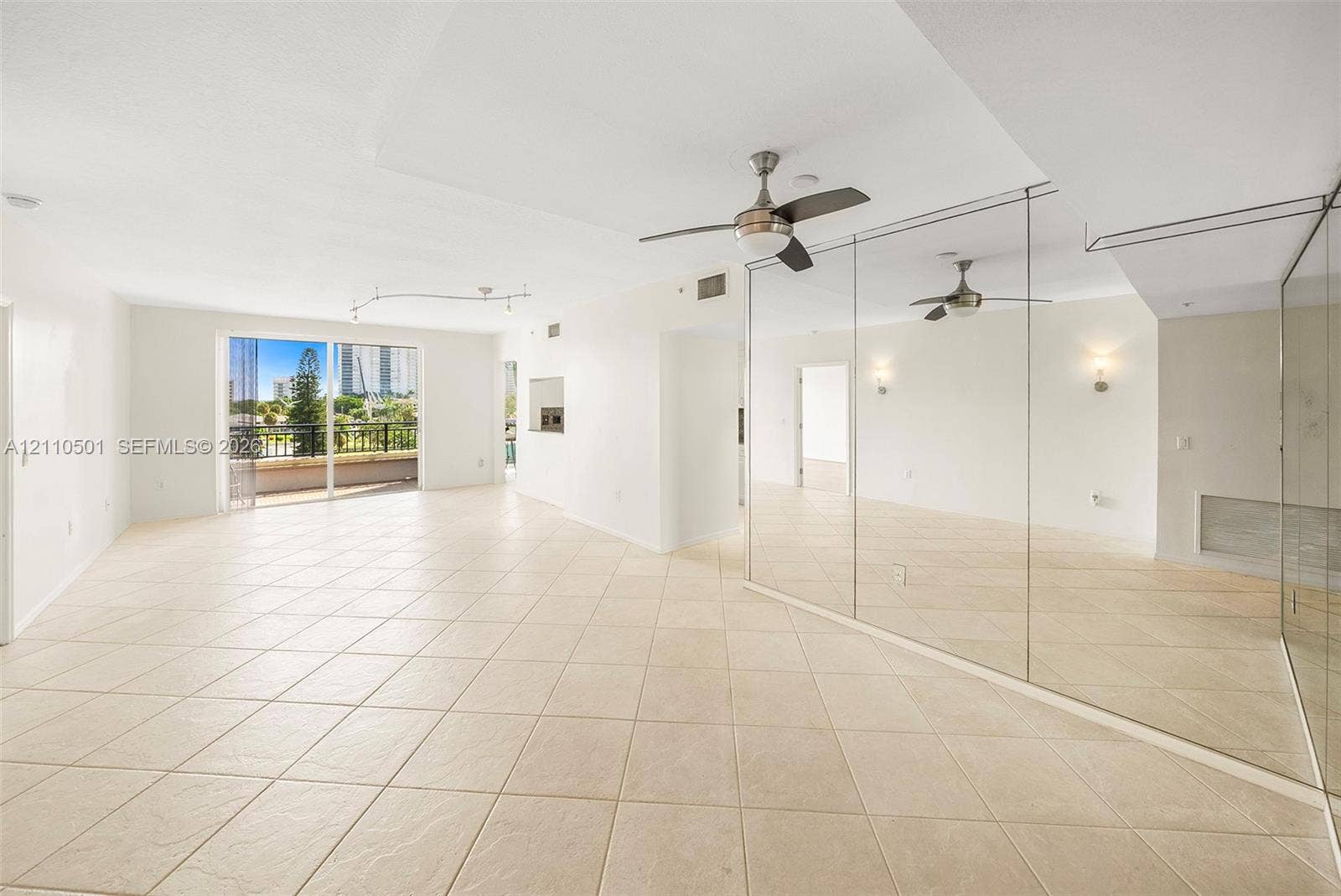 Open interior space featuring neutral tile flooring, floor-to-ceiling wall mirrors, a ceiling fan with integrated lighting, and a sliding glass door opening to an exterior balcony