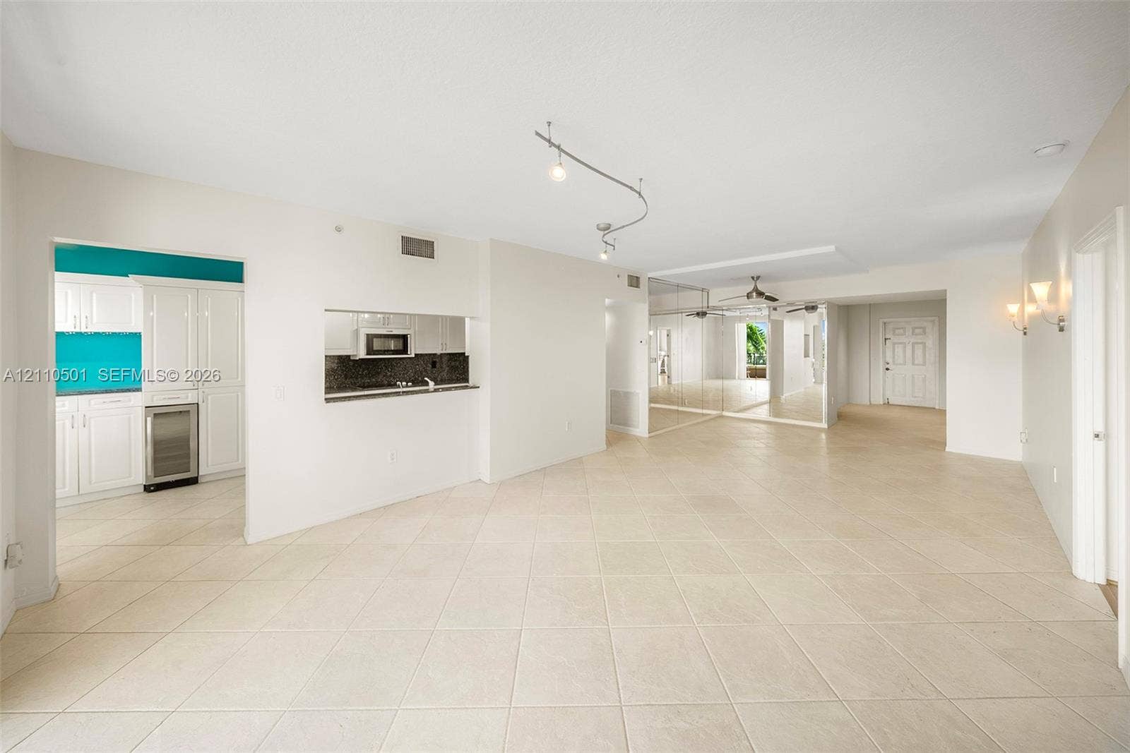 Open-concept interior featuring neutral tile flooring, track lighting, mirrored wall accents, and a kitchen service window with dark stone countertops