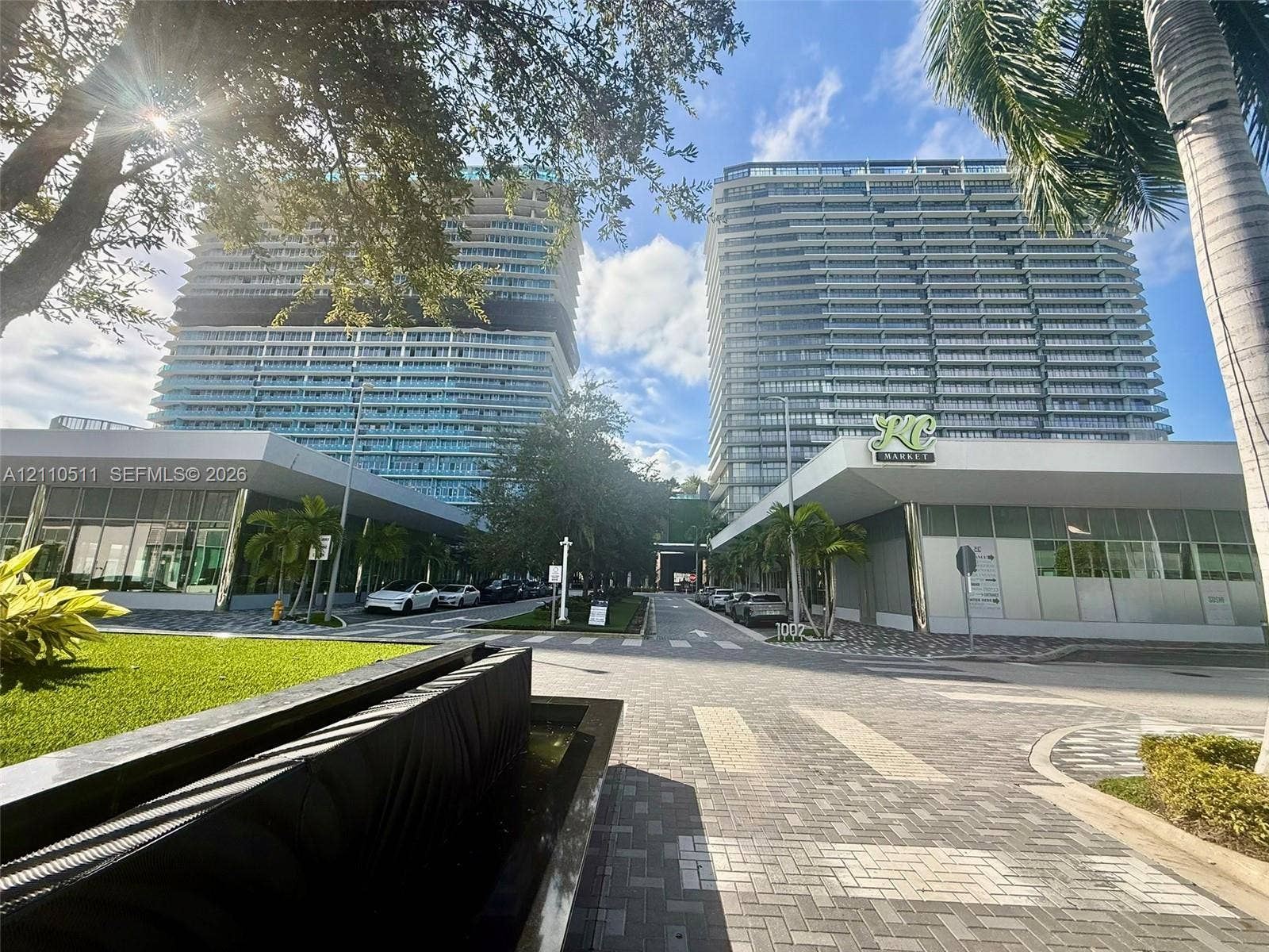 1170 E Hallandale Beach Boulevard, #1208, Hallandale Beach, FL 33009 Modern high-rise towers featuring floor-to-ceiling glass facades, covered ground-level commercial spaces, paved stone driveways, and landscaped green areas