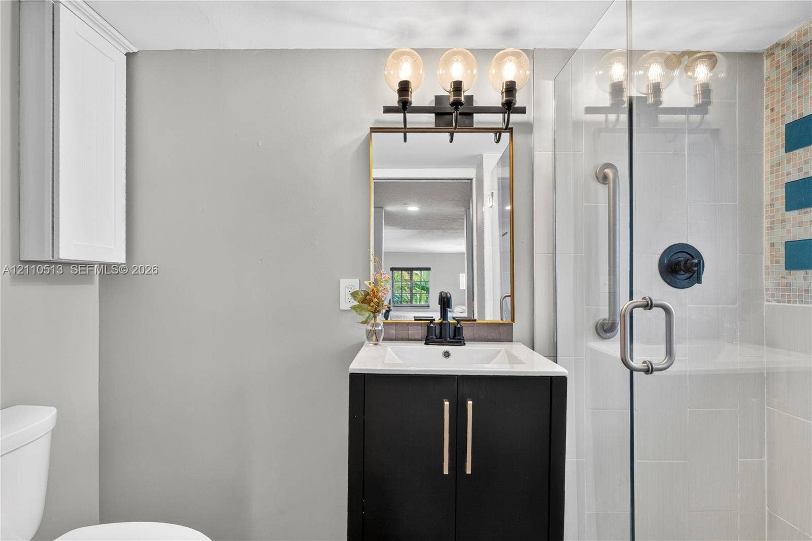 6520 SW 16th Terrace, West Miami, FL 33155 Bathroom featuring a vanity with dark cabinetry and a light-toned countertop, three-light fixture, gold-framed mirror, and a glass-enclosed shower with tiled surround
