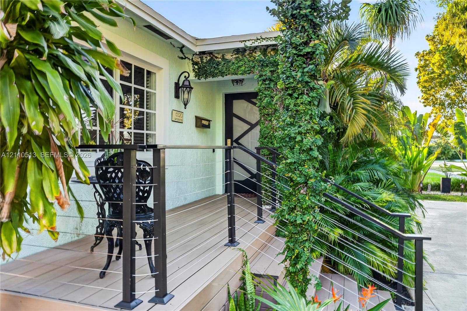 6520 SW 16th Terrace, West Miami, FL 33155 Elevated porch featuring composite decking, black metal cable railings, custom geometric front door, and traditional wall-mounted lantern lighting