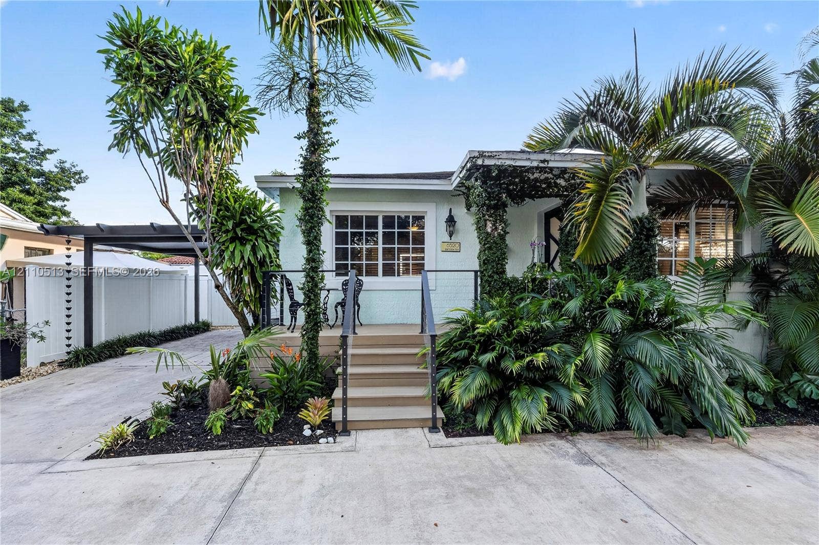 6520 SW 16th Terrace, West Miami, FL 33155 Light-colored exterior siding with grid-pattern windows, raised wood-finish front porch, black metal railing, concrete driveway, and covered carport