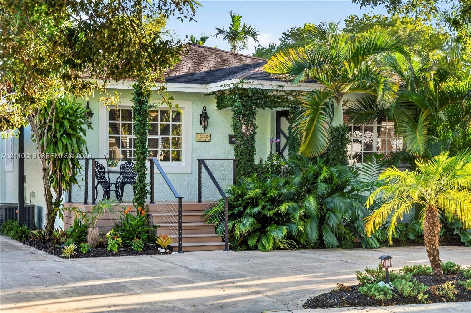 6520 SW 16th Terrace, West Miami, FL 33155 Exterior facade featuring a covered porch with cable railing, shingle roof, multi-pane windows, and extensive mature tropical landscaping