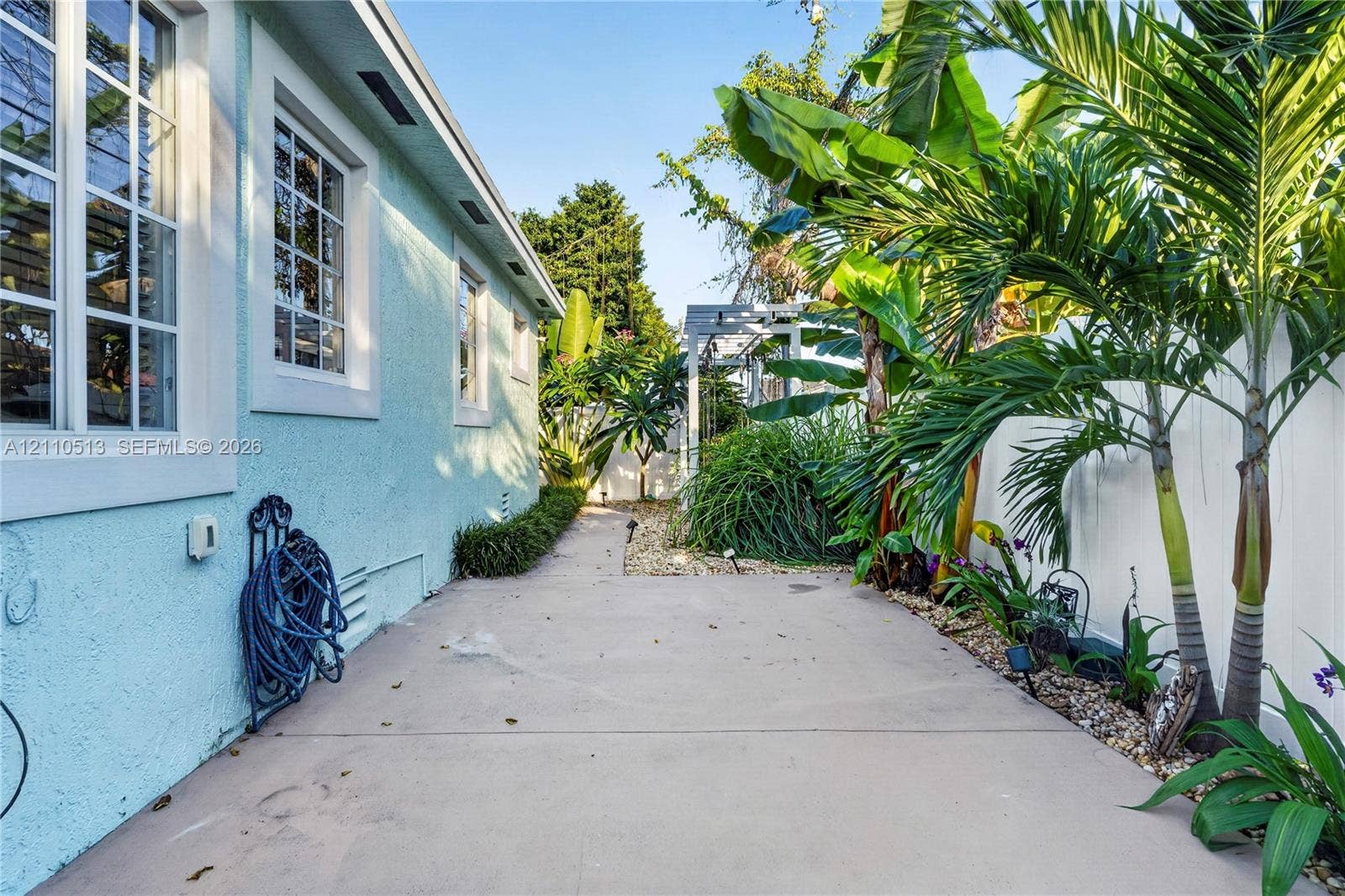 6520 SW 16th Terrace, West Miami, FL 33155 Exterior side yard featuring a concrete walkway, light blue stucco wall, multi-pane windows, privacy fencing, and established tropical landscaping