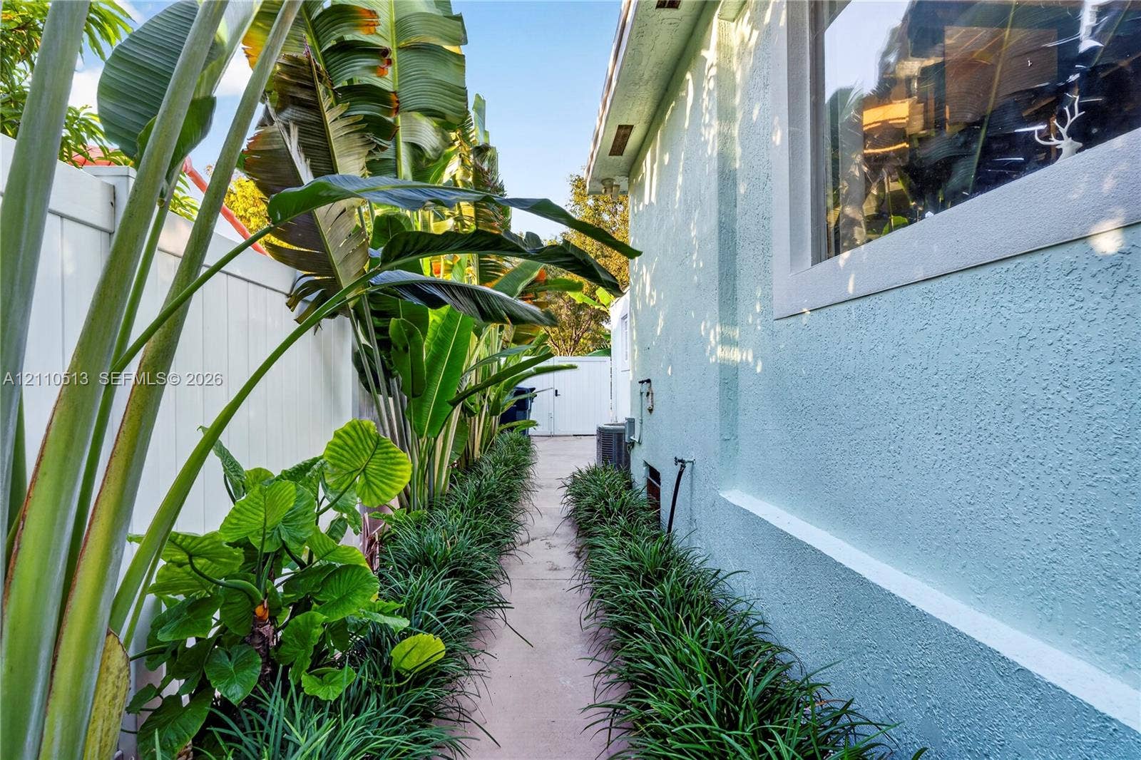 6520 SW 16th Terrace, West Miami, FL 33155 Side yard featuring a concrete pathway, privacy vinyl fencing, mature tropical landscaping, and textured stucco exterior wall