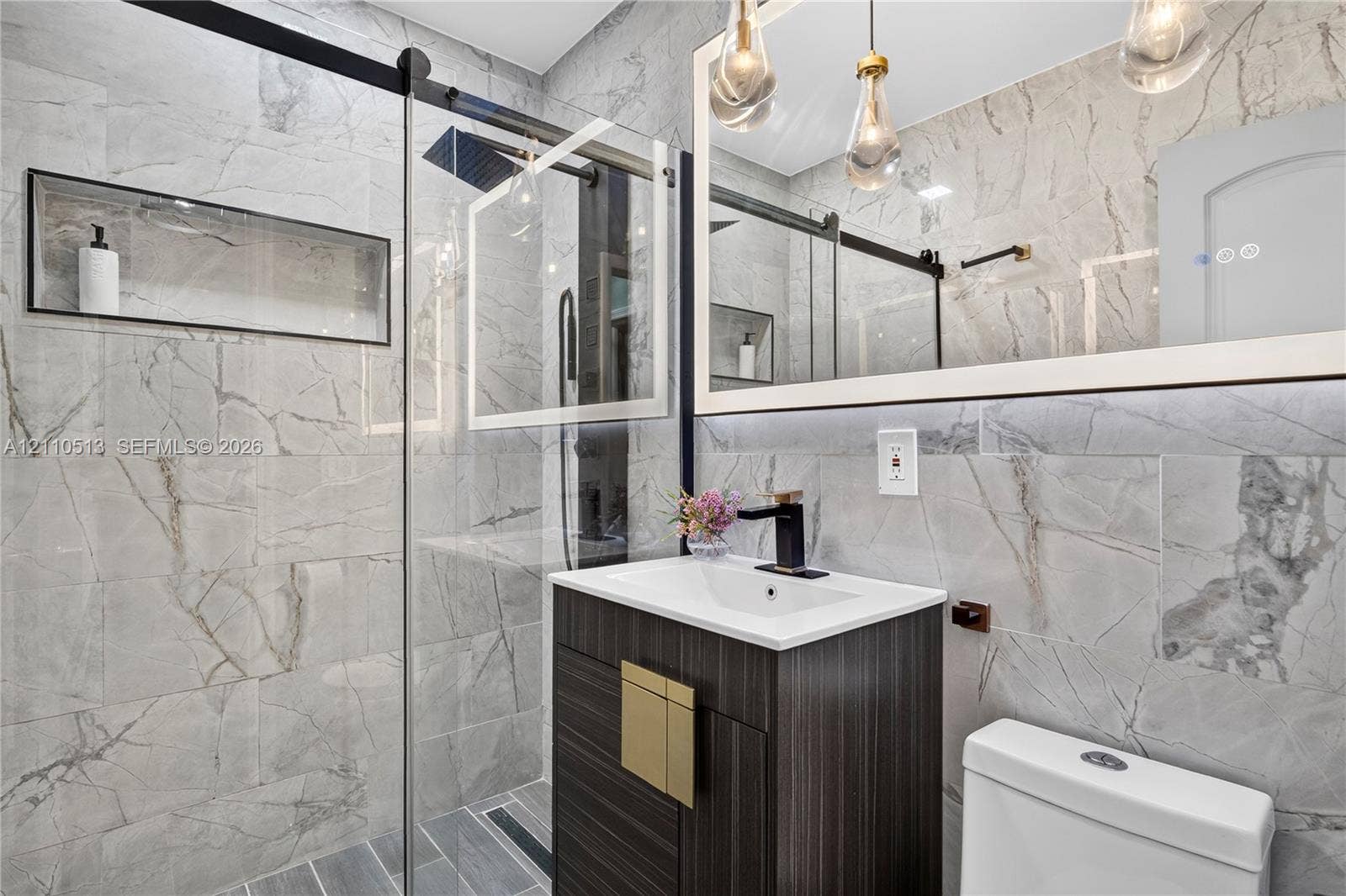 6520 SW 16th Terrace, West Miami, FL 33155 Bathroom featuring full-height large-format stone tile, a glass-enclosed shower with a built-in recessed wall niche, a dark wood-finish vanity with a rectangular sink, matte black fixtures, and a framed mirror with pendant lighting