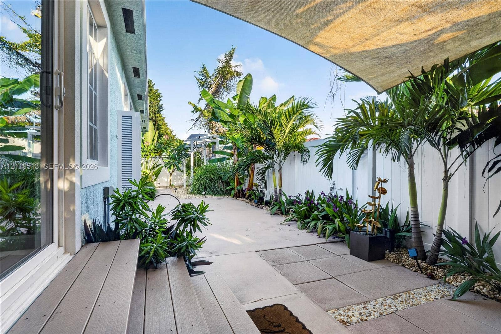 6520 SW 16th Terrace, West Miami, FL 33155 Rear patio featuring a concrete and paved surface, wood-finish decking steps, a retractable shade canopy, white privacy fencing, and mature tropical landscaping