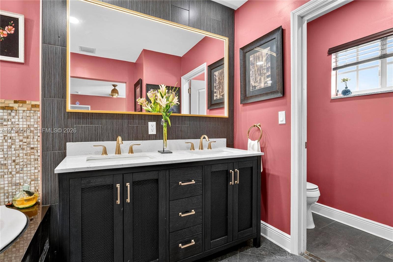 6520 SW 16th Terrace, West Miami, FL 33155 Double vanity featuring a white countertop, dual undermount sinks, gold-tone hardware, and an expansive wall-mounted mirror framed in gold, set against a dark tiled backsplash
