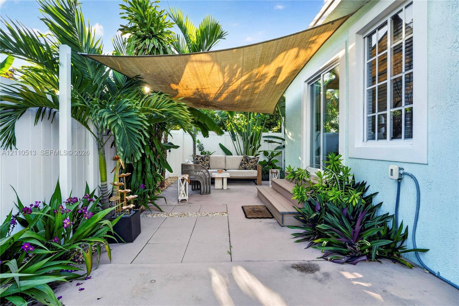 6520 SW 16th Terrace, West Miami, FL 33155 Paved patio area featuring a mounted shade sail, wood-finish stepped platform, light-colored stucco siding, and surrounding privacy fencing