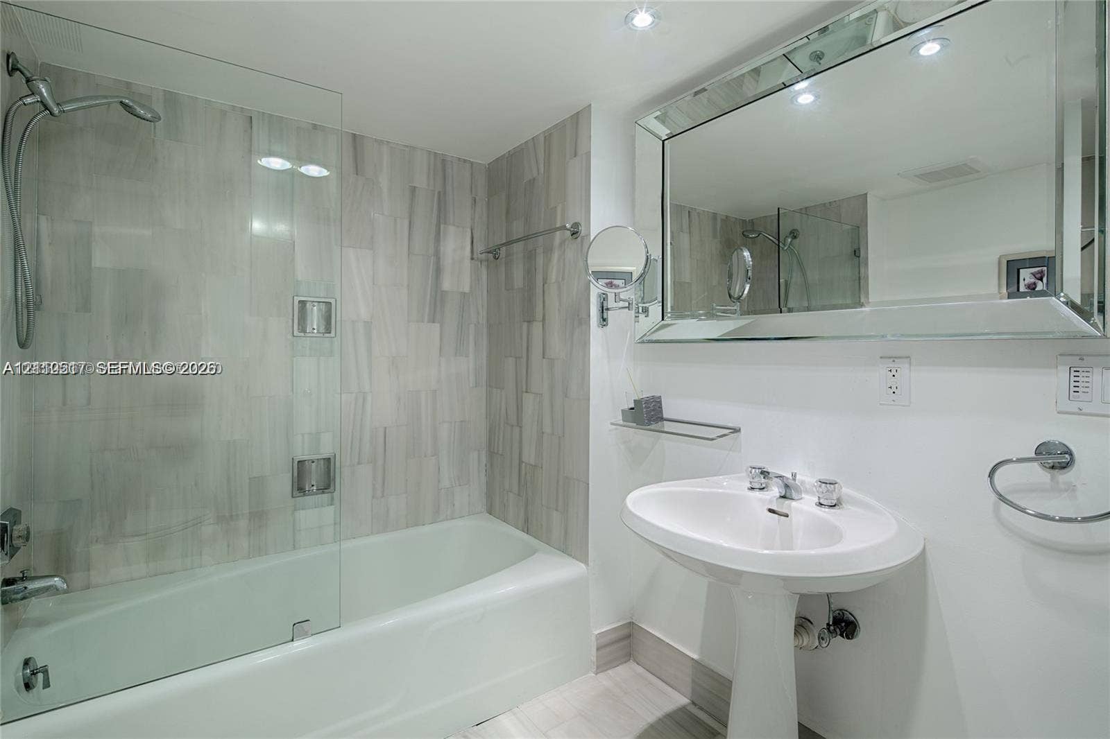 Bathroom featuring a white pedestal sink, frameless glass shower screen, tub with wall-mounted shower head, large beveled mirror, and stone-finish wall tile
