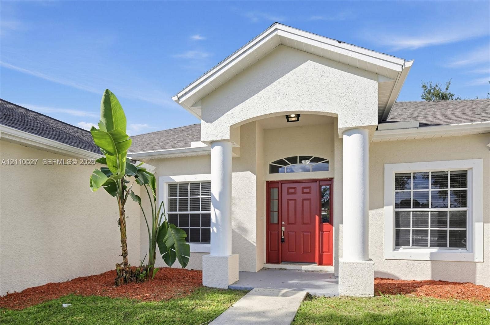 5509 NW Commodore Terrace, Port St Lucie, FL 34983 Stucco exterior featuring an arched entryway with decorative columns, a prominent red entry door with sidelights, and a transom window