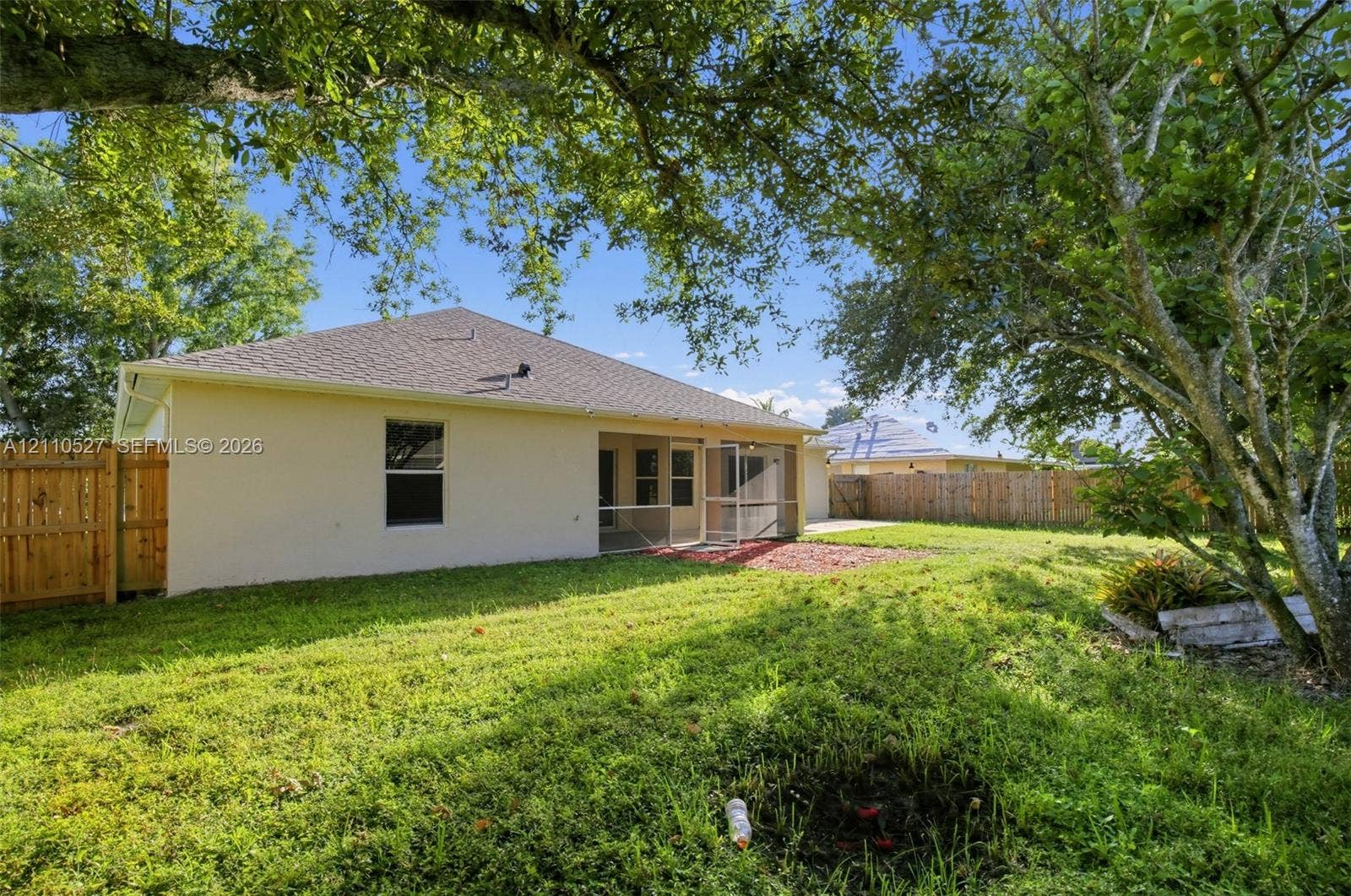5509 NW Commodore Terrace, Port St Lucie, FL 34983 Rear exterior featuring stucco siding, a screened-in lanai, wood privacy fencing, and mature trees