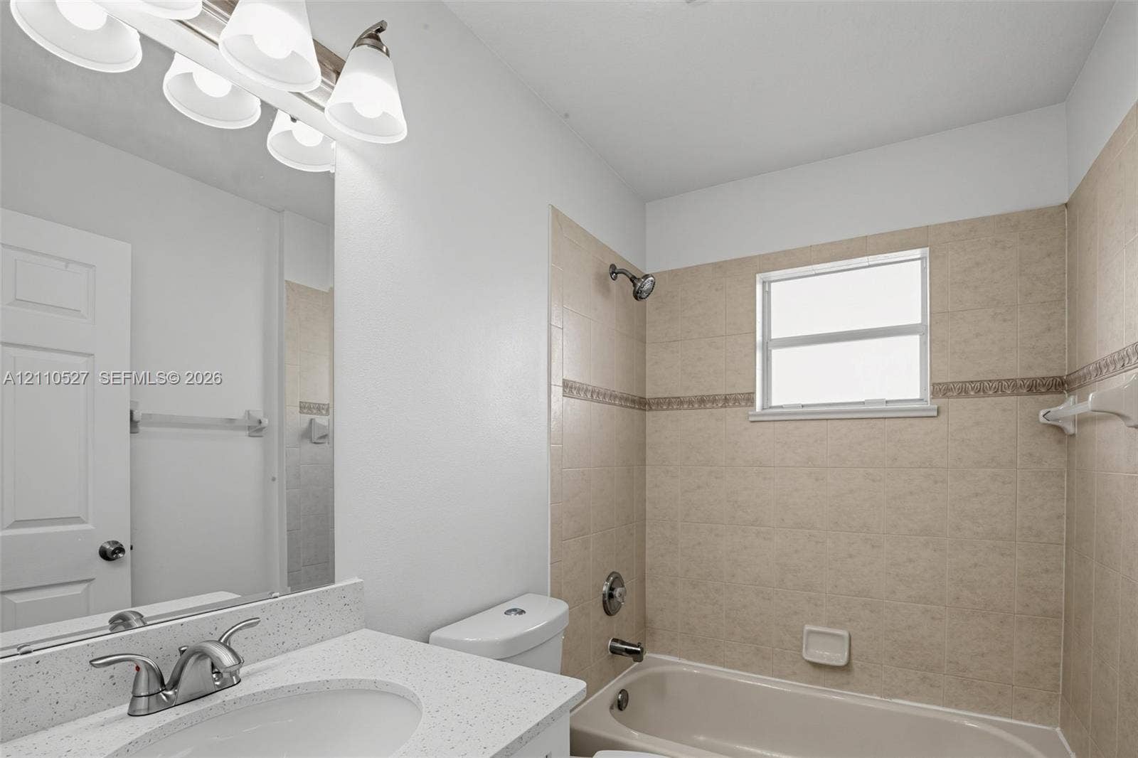 5509 NW Commodore Terrace, Port St Lucie, FL 34983 Bathroom featuring a speckled countertop with an integrated basin, multi-bulb vanity light, polished chrome hardware, and neutral tile surround with a decorative border accent