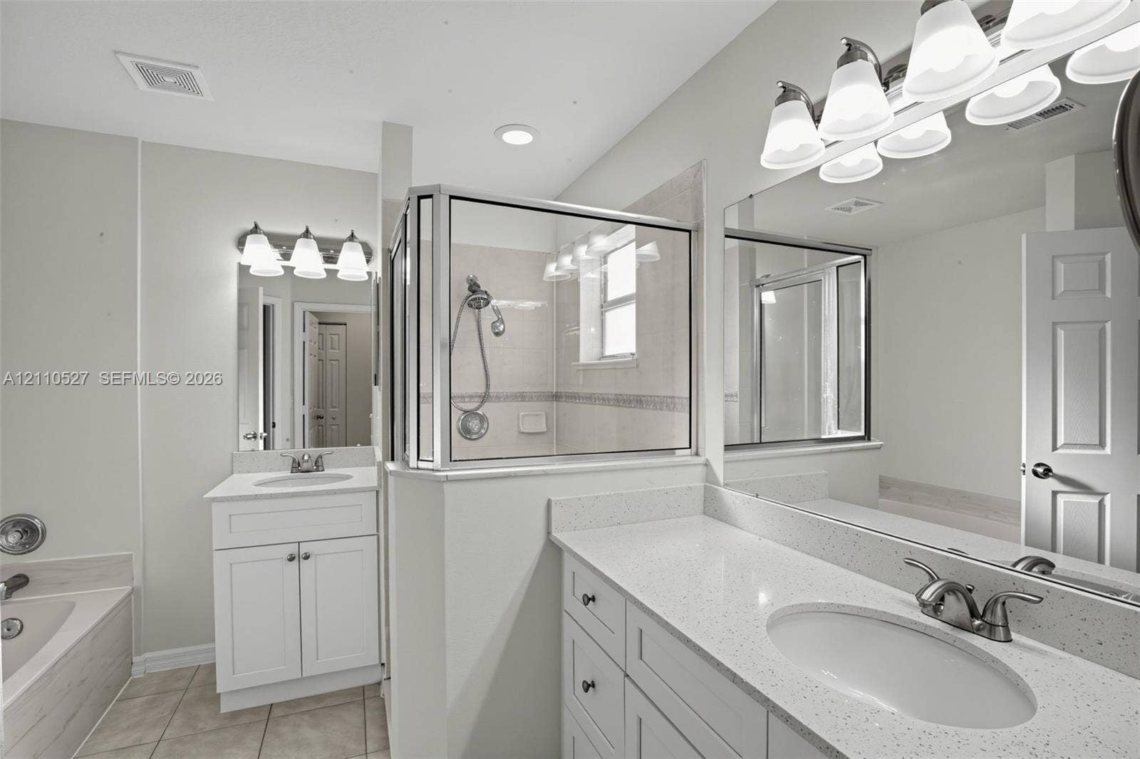 5509 NW Commodore Terrace, Port St Lucie, FL 34983 Bathroom featuring dual white shaker-style cabinetry, light-toned stone-finish countertops, an enclosed glass-paneled shower with tiled surround, and large-scale vanity mirrors with multi-bulb light fixtures