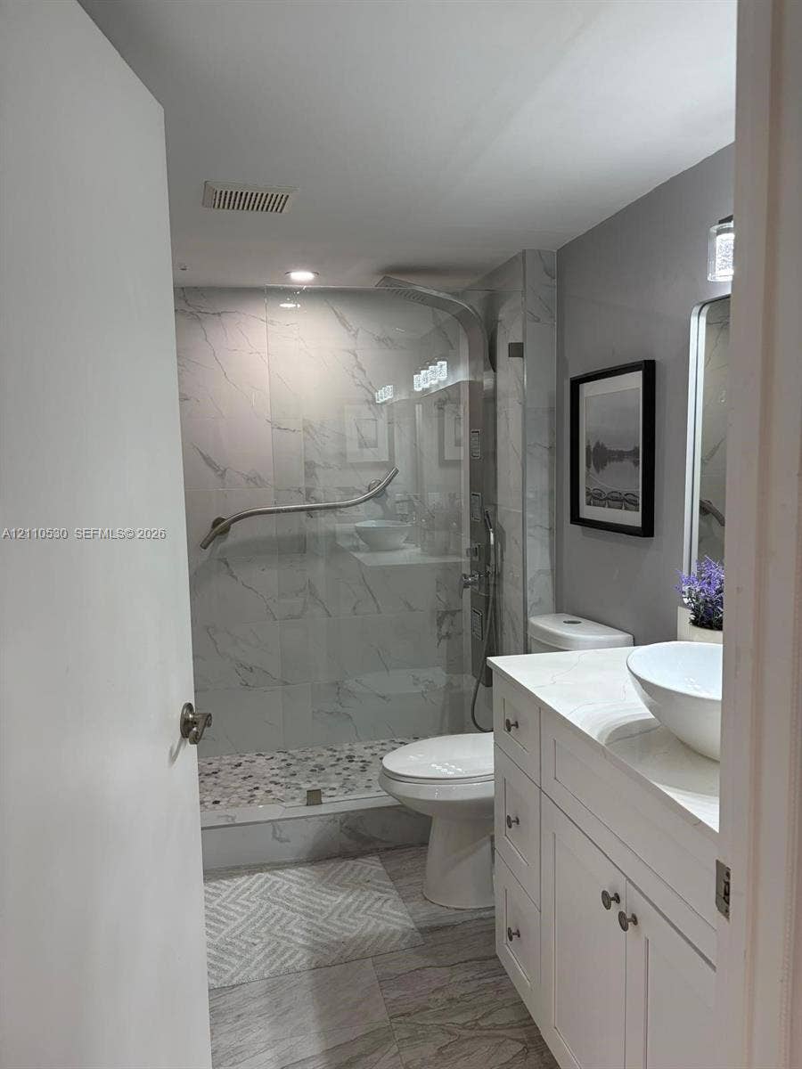13255 SW 16th Court, #K112, Pembroke Pines, FL 33027 Bathroom featuring a white vanity cabinet with an integrated white vessel sink, walk-in shower with stone-tile flooring and glass enclosure, and stone-finish wall tile