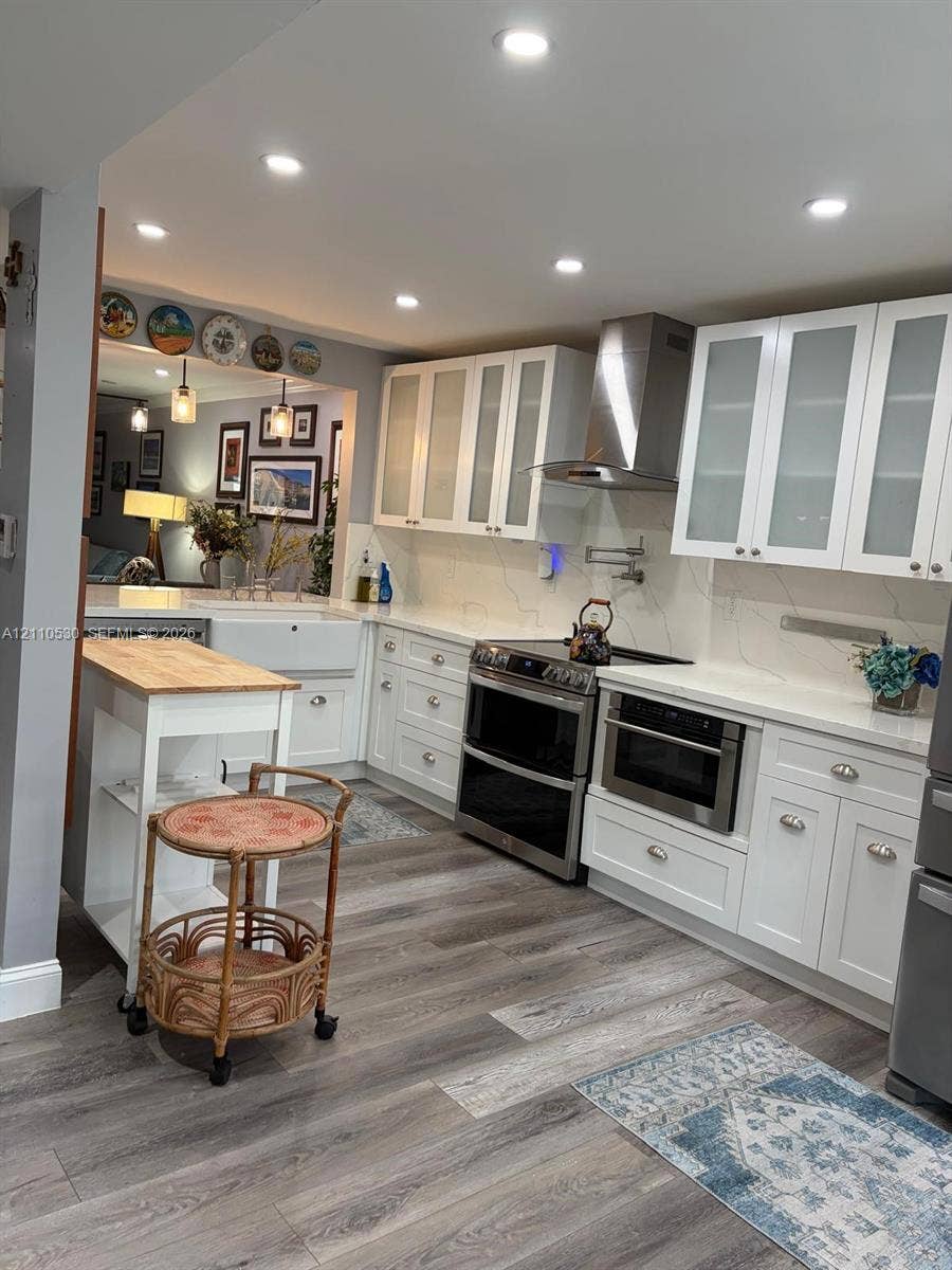 13255 SW 16th Court, #K112, Pembroke Pines, FL 33027 Kitchen featuring white cabinetry with glass-front uppers, stone-finish countertops, apron-front sink, stainless steel range hood, and wood-finish flooring
