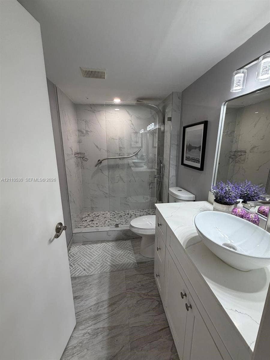13255 SW 16th Court, #K112, Pembroke Pines, FL 33027 Bathroom featuring a white vanity with a rectangular vessel sink and solid-surface countertop, walk-in shower with stone-patterned wall tile and pebble-texture flooring, and large-format tile flooring