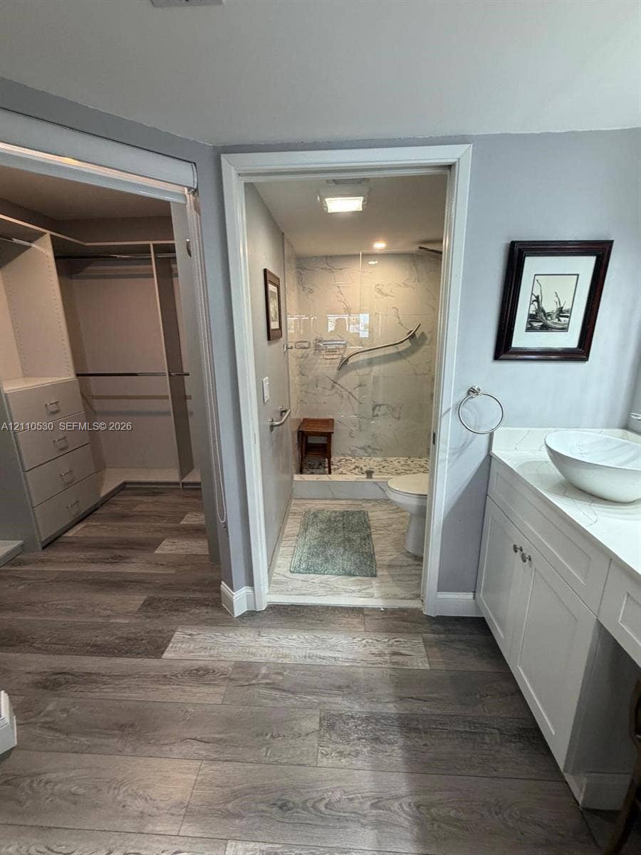 13255 SW 16th Court, #K112, Pembroke Pines, FL 33027 Bathroom featuring a white vanity with a vessel sink, wood-finish flooring, and a walk-in shower with stone-look wall tile and pebble-tile flooring, paired with a custom-built walk-in closet