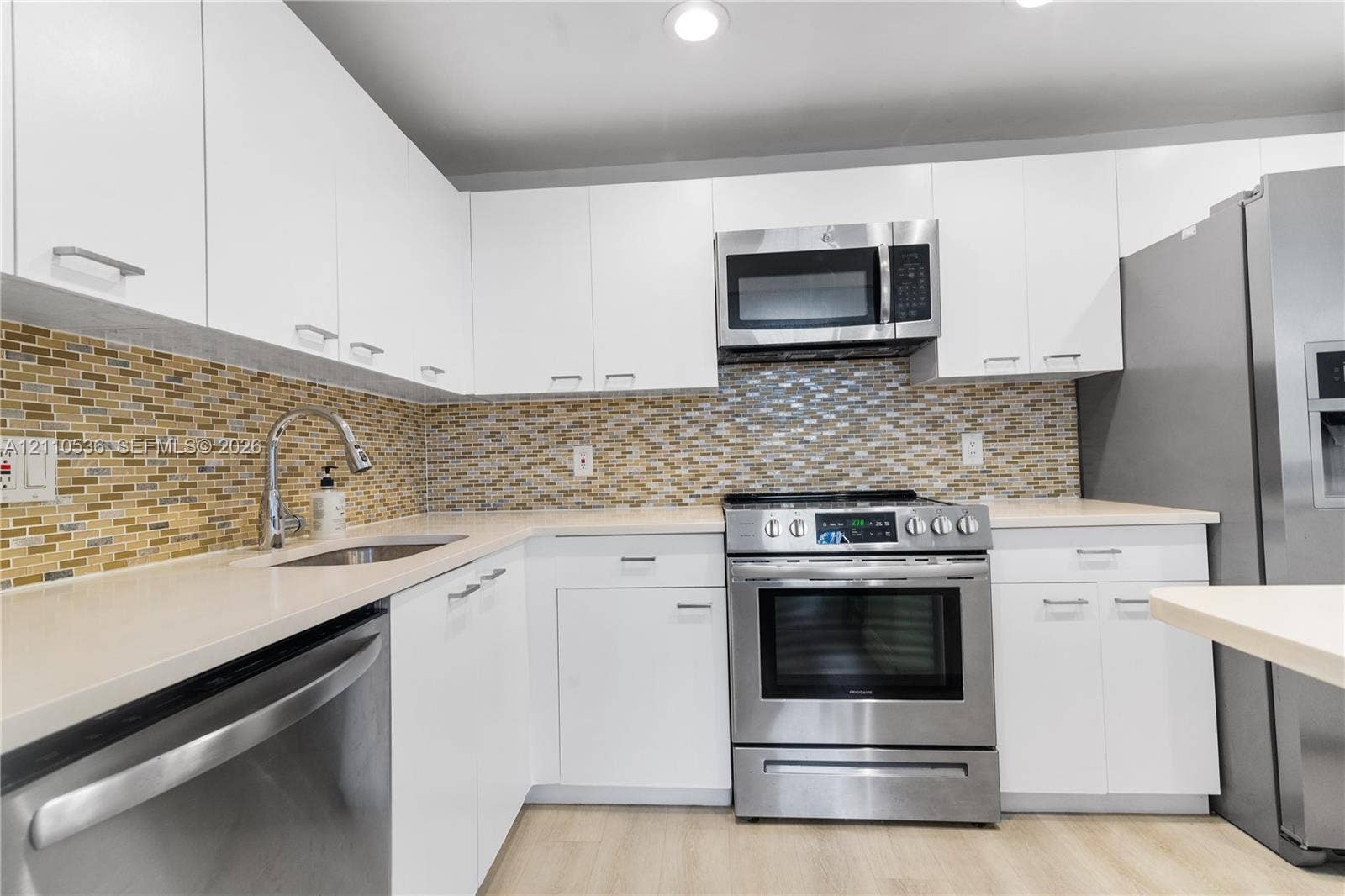 Kitchen featuring flat-panel white cabinetry, stainless steel appliances, multi-tonal mosaic tile backsplash, solid-surface countertops, and wood-finish flooring
