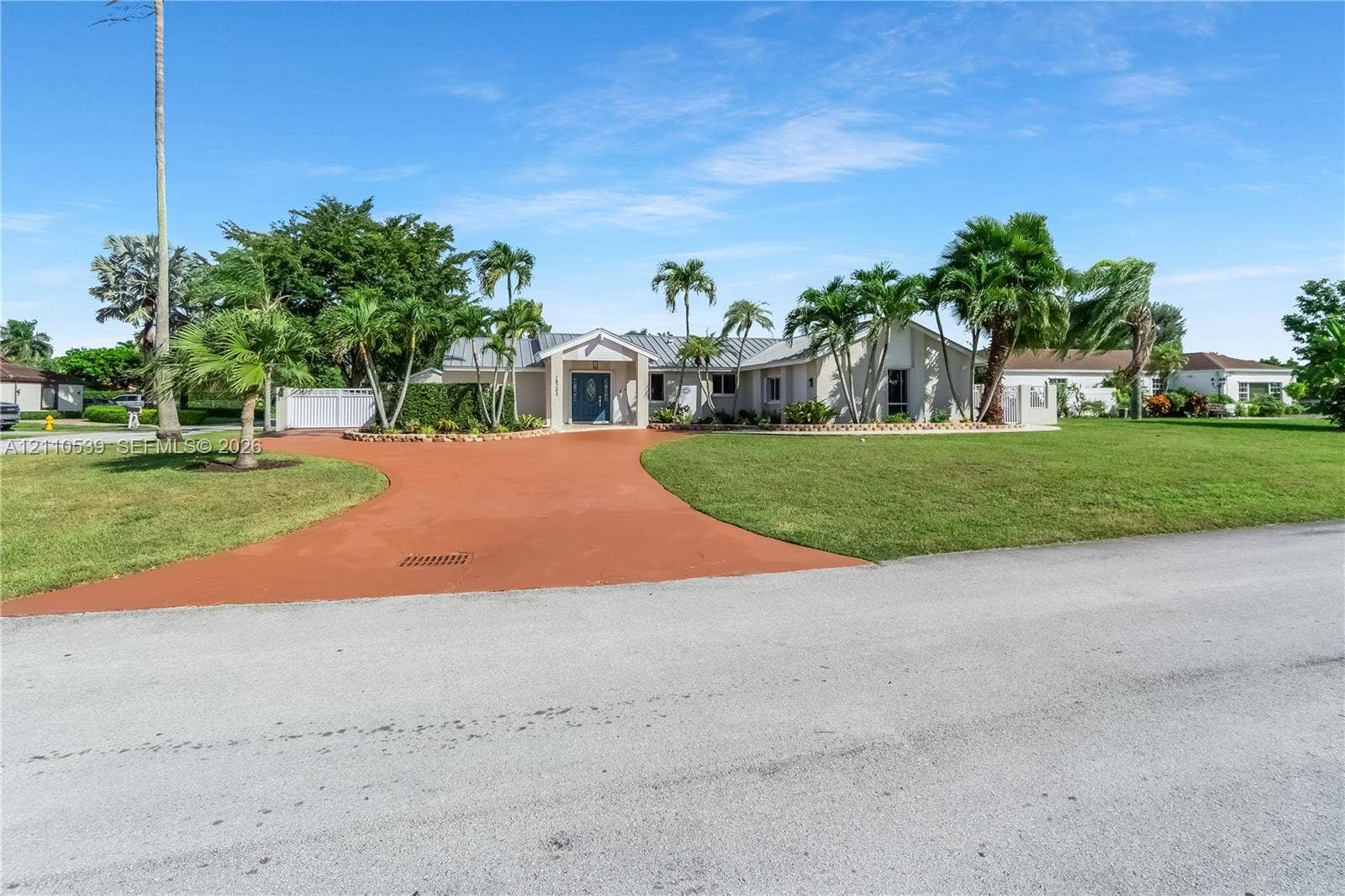 Single-story residence featuring a metal roof, an expansive stained concrete driveway, mature palm trees, and a gated side perimeter
