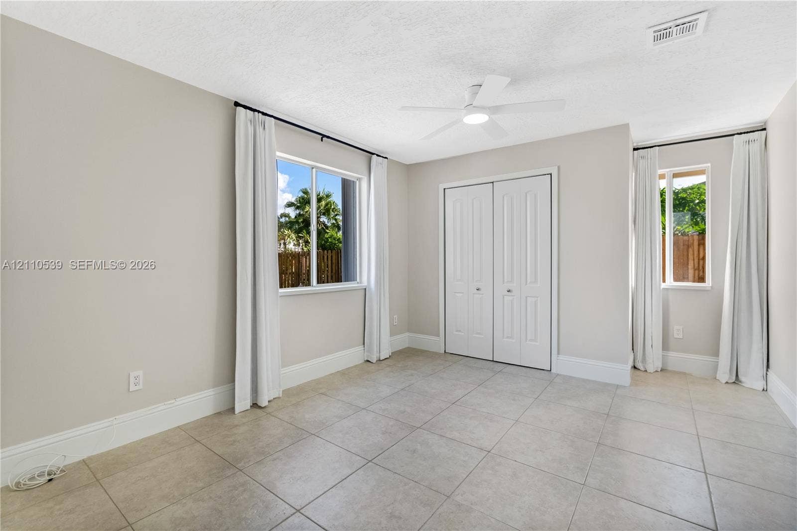 Neutral-toned interior featuring large-format ceramic tile flooring, dual-aspect windows, ceiling-mounted paddle fan, and double-door closet cabinetry