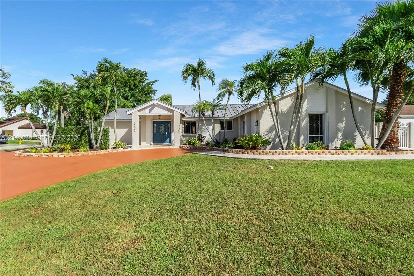 Single-story residence featuring a painted stucco exterior, metal roofing, expansive front lawn, terracotta-toned driveway, and manicured palm landscaping