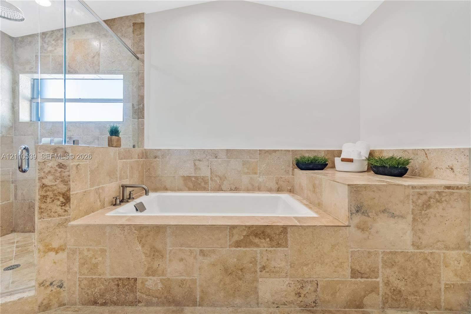 Built-in soaking tub with natural stone tile surround, integrated tiled ledge, and glass-enclosed shower with metallic hardware