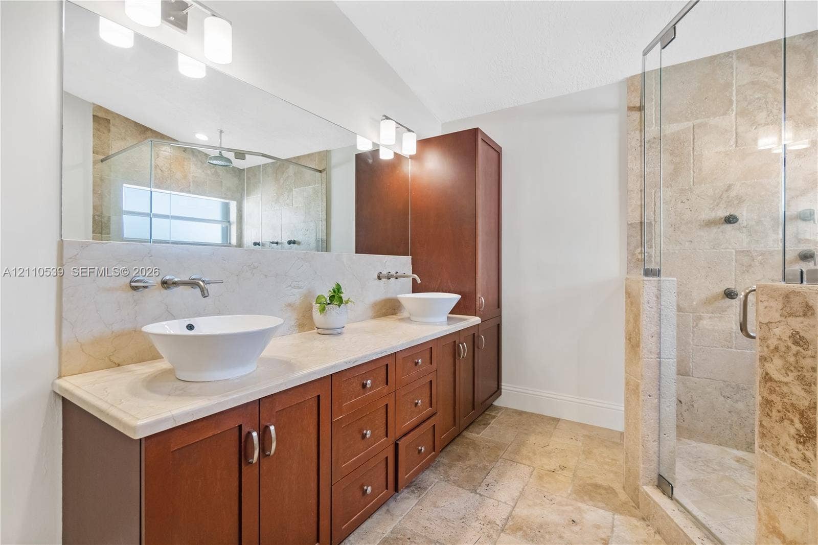 Dual vessel sinks featuring wall-mounted faucets, a stone-topped vanity with built-in cabinetry, large-format mirror, and a walk-in shower with stone tile surround