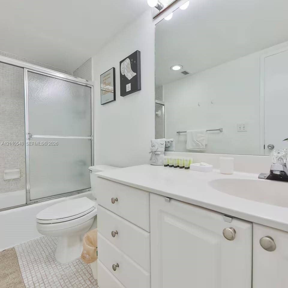 Bathroom featuring a white vanity with integrated basin, polished metal hardware, a wall-spanning mirror, frosted glass sliding shower enclosure, and mosaic floor tile