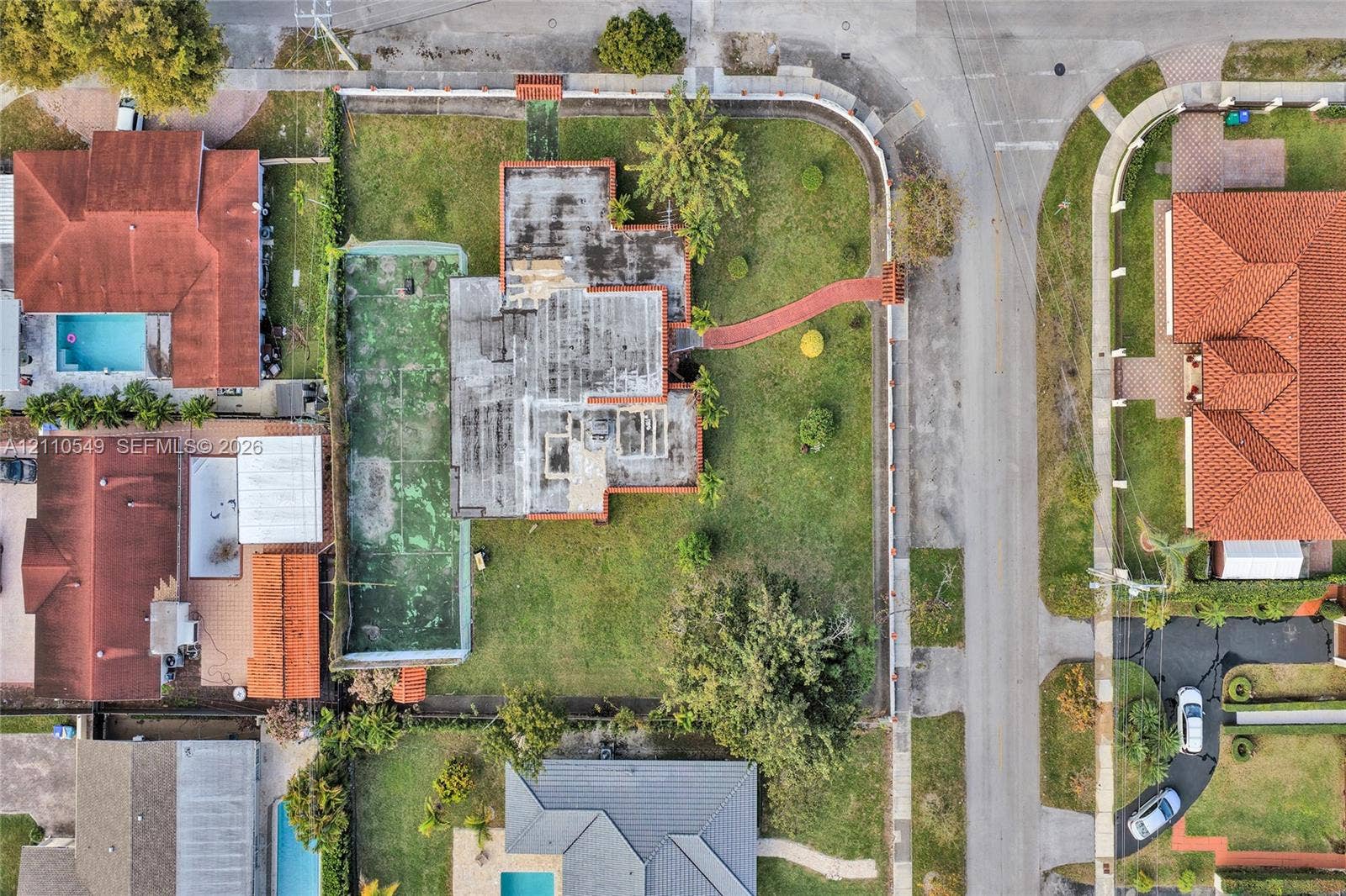 500 SW 39th Court, Miami, FL 33134 Aerial perspective of a corner lot property featuring a private tennis court, expansive lawn, perimeter masonry wall, and paved walkway