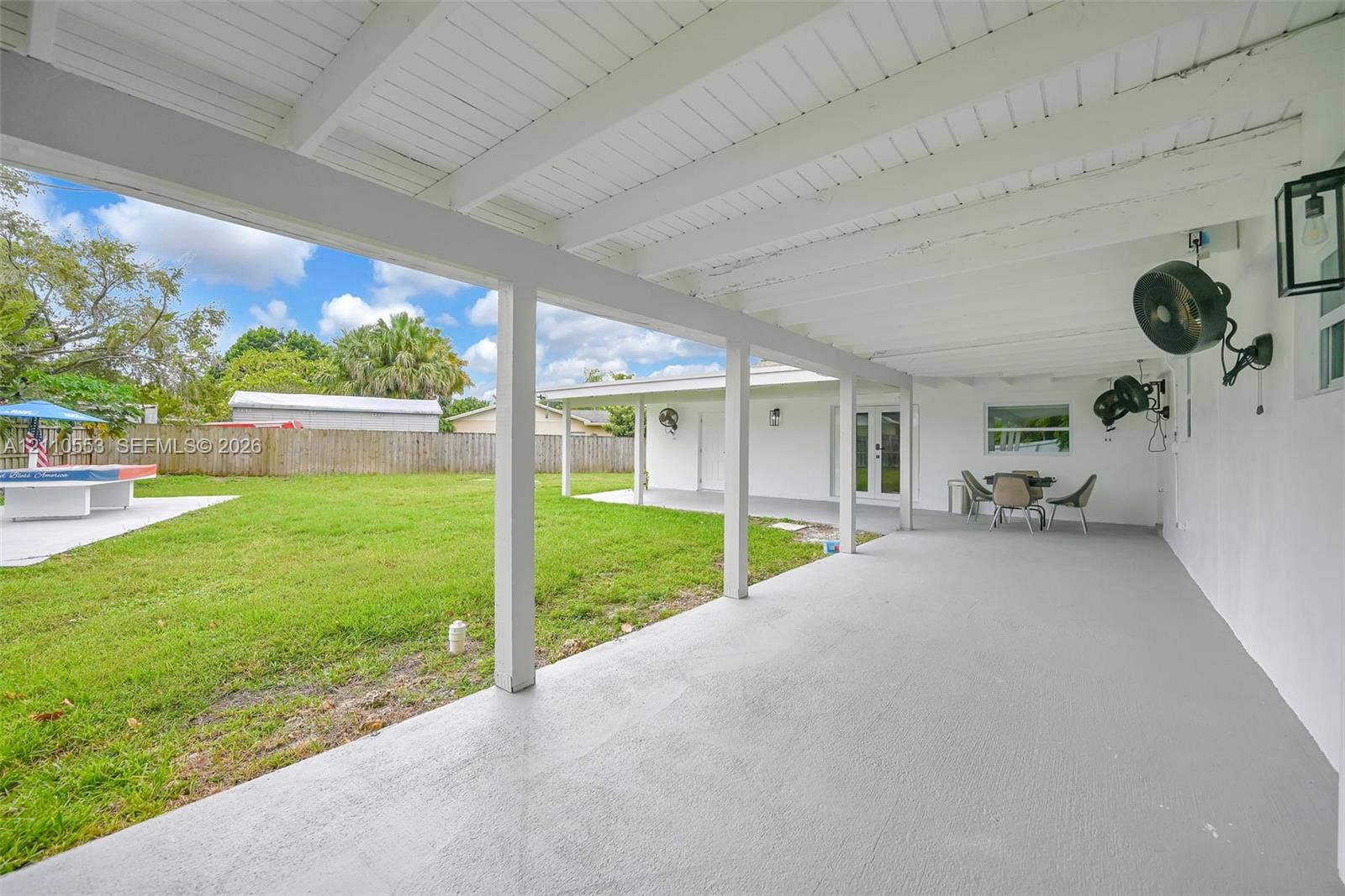 16611 SW 108th Court, Miami, FL 33157 Covered rear patio featuring a painted concrete floor, wood-beam ceiling, wall-mounted outdoor lighting, and expansive backyard space enclosed by wood fencing