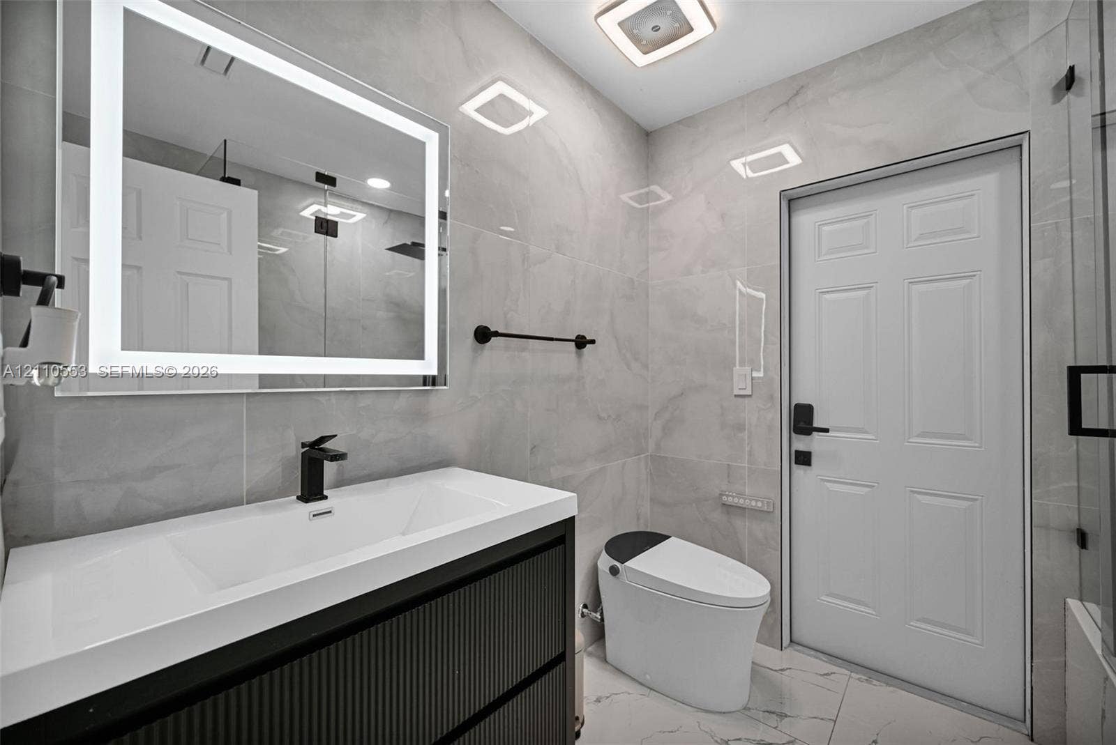 16611 SW 108th Court, Miami, FL 33157 Bathroom featuring a modern vanity with a rectangular integrated sink, wall-mounted matte black faucet, illuminated LED mirror, floor-to-ceiling large-format tile, and a glass-enclosed shower