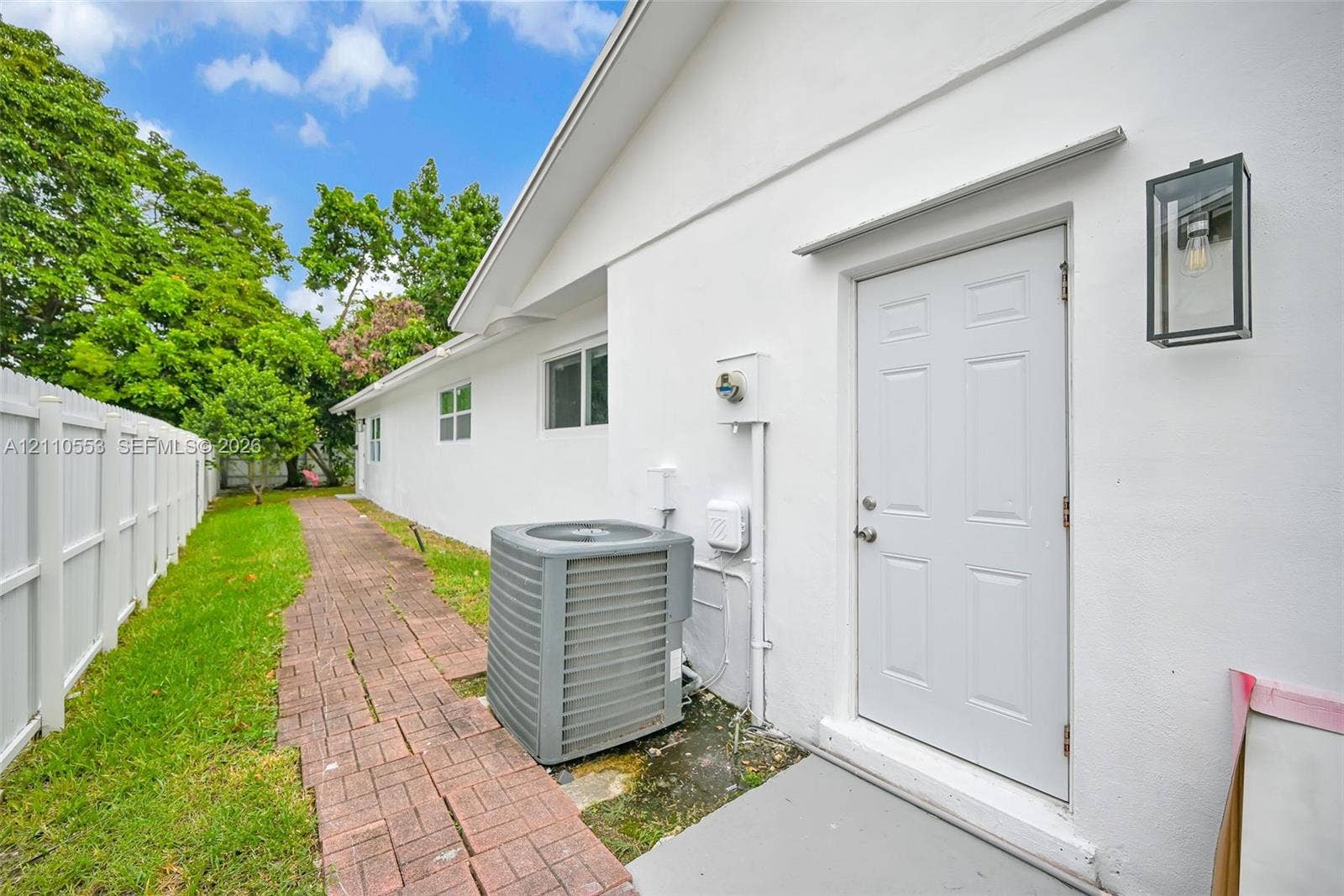16611 SW 108th Court, Miami, FL 33157 Exterior side profile featuring a white stucco finish, a paved brick walkway, a white vinyl privacy fence, and a modern black-framed wall-mounted lantern