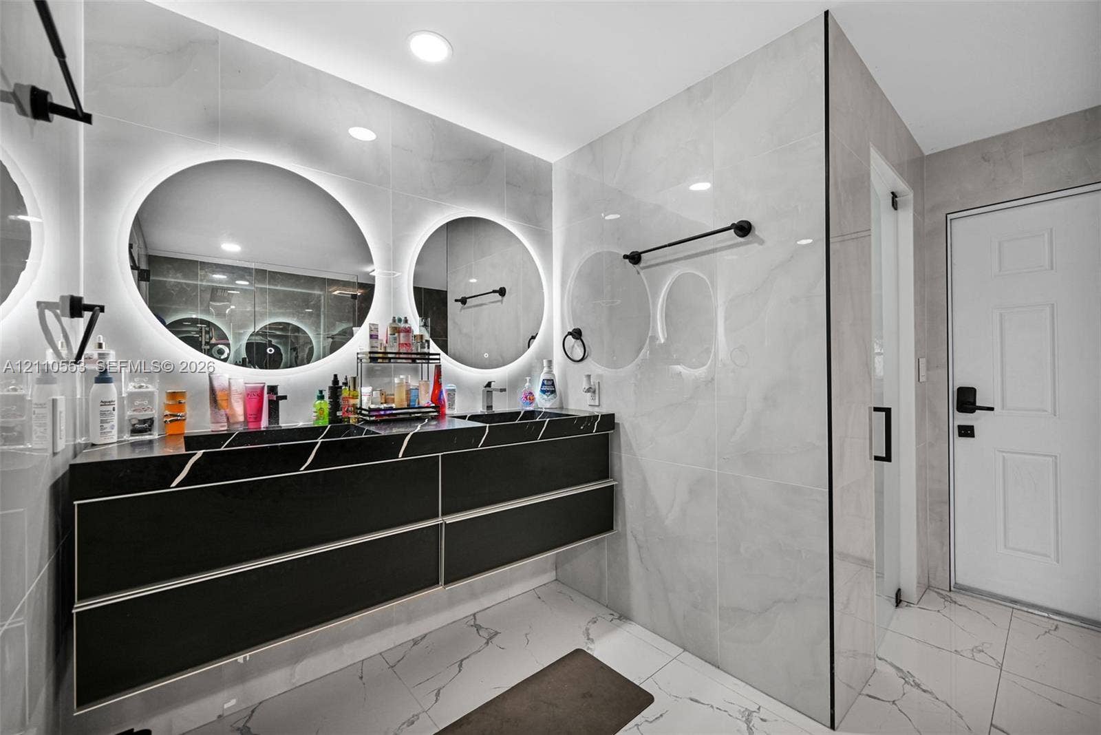 16611 SW 108th Court, Miami, FL 33157 Floating dual-sink vanity with matte black finish, integrated backlit circular mirrors, large-format polished wall tile, and light-toned floor tile with dark veining