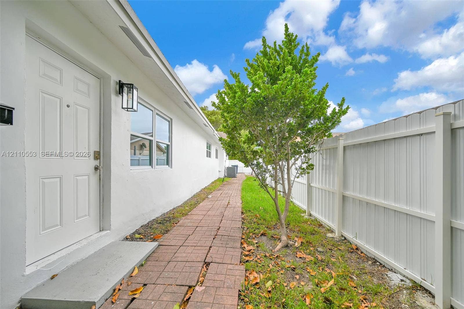16611 SW 108th Court, Miami, FL 33157 Exterior side path featuring a brick paver walkway, white smooth-stucco siding, a white paneled door, black metal wall-mounted lighting, and a white privacy fence