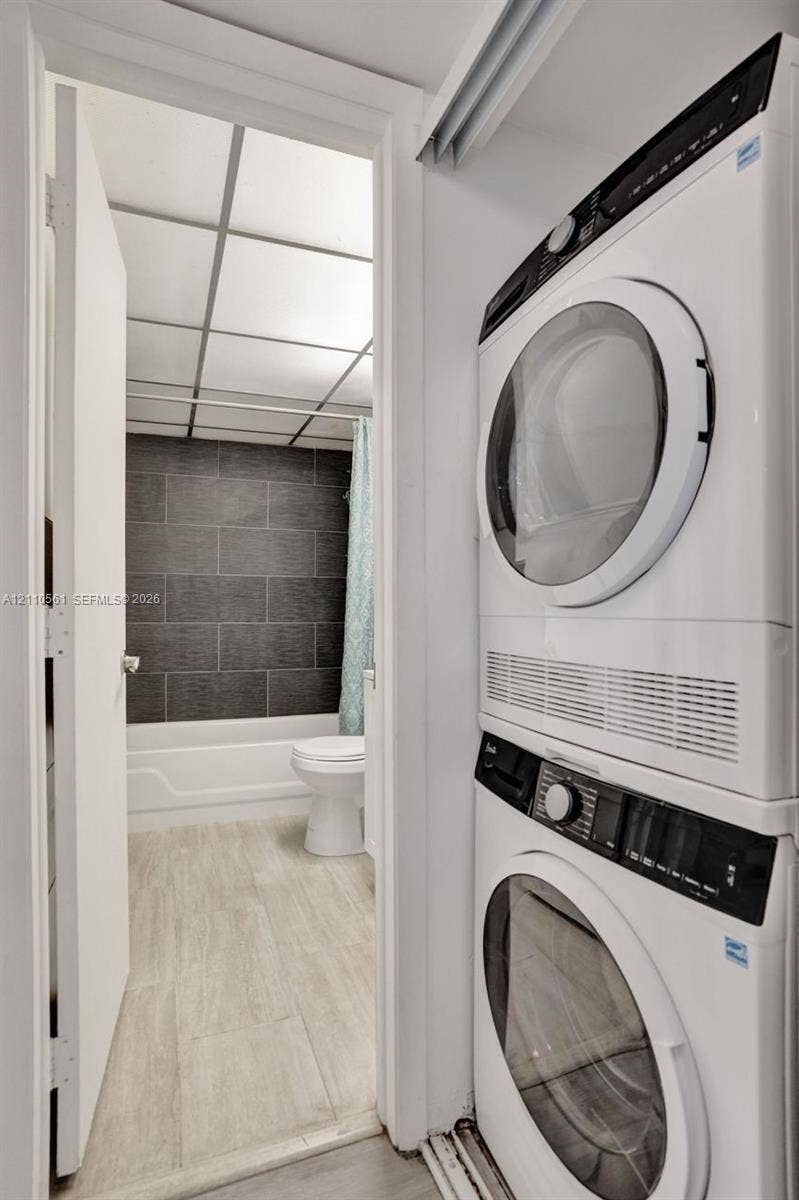 7950 W Mcnab Road, #222, Tamarac, FL 33321 Stacked laundry unit featuring a front-loading washer and dryer, situated near a bathroom with wood-finish flooring, a white bathtub, and dark horizontal wall tiling