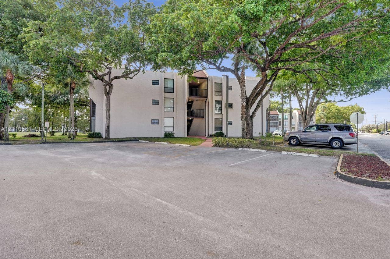 7950 W Mcnab Road, #222, Tamarac, FL 33321 Multi-story building with light-toned facade, asphalt parking area, and mature canopy trees