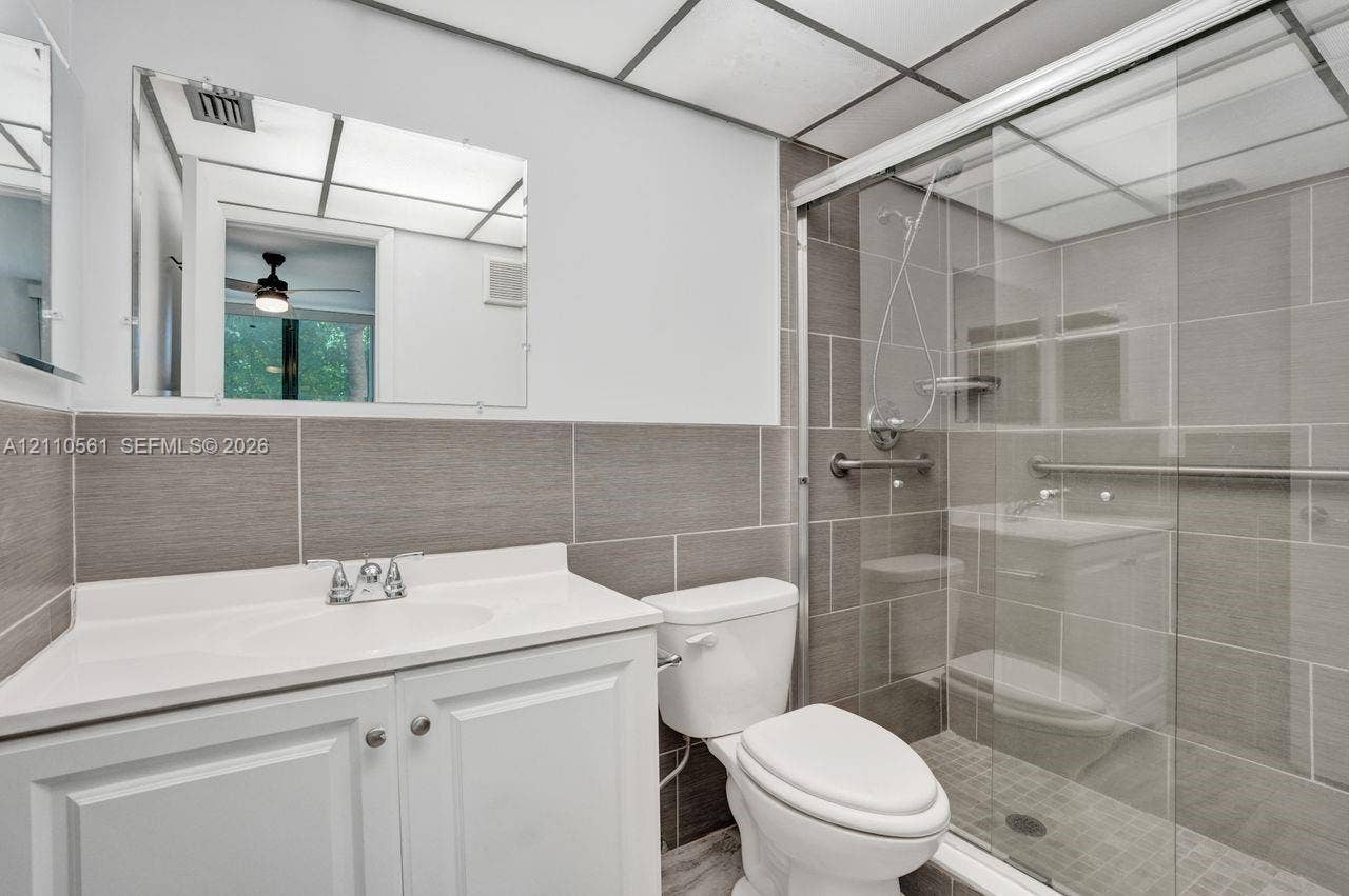 7950 W Mcnab Road, #222, Tamarac, FL 33321 Bathroom featuring a white vanity cabinet with an integrated sink, rectangular tile surround, glass-enclosed shower, and grid-patterned ceiling panels