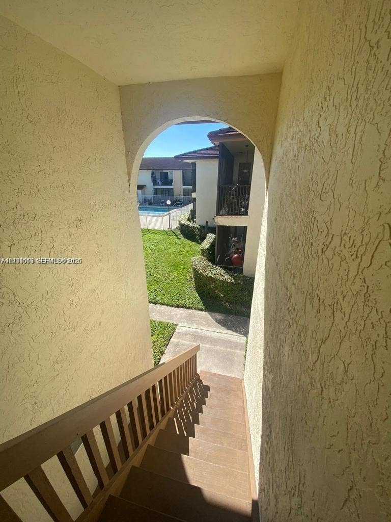 Exterior staircase featuring stucco walls, an arched entryway, and wooden handrail, with grounds displaying manicured landscaping and a swimming pool