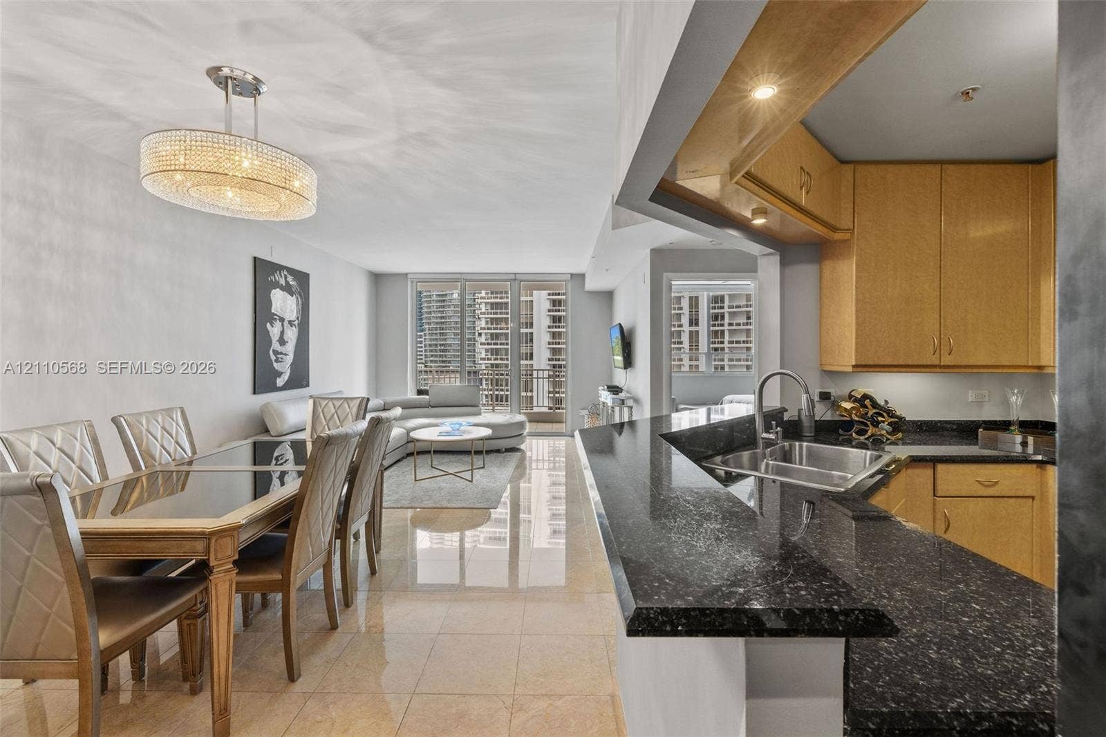 801 Brickell Key Boulevard, #2006, Miami, FL 33131 Open-concept living space featuring high-gloss tile flooring, kitchen with dark stone-finish countertops, custom light-toned wood cabinetry, stainless steel undermount sink, and a multi-tier crystal chandelier