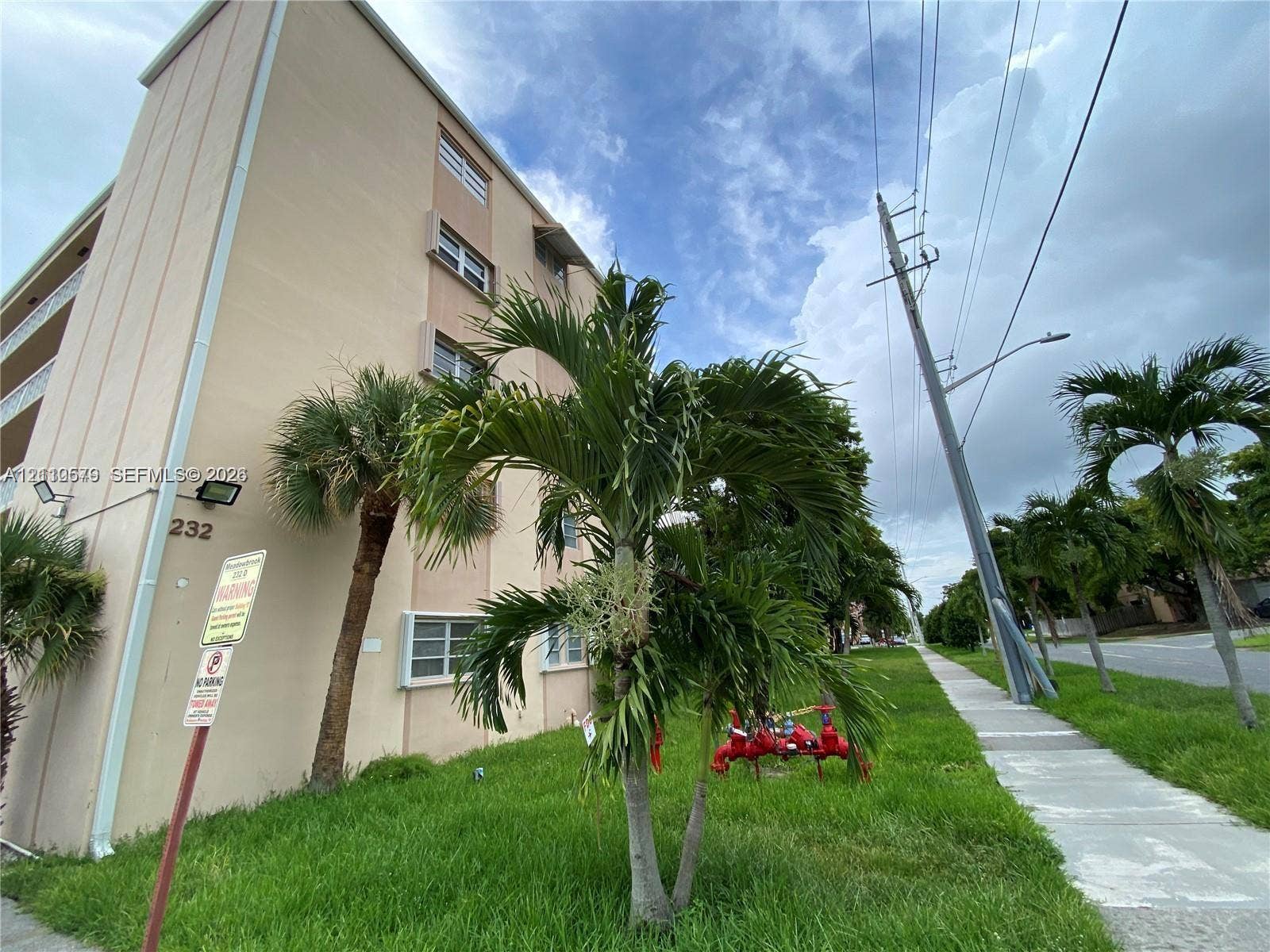 232 NE 12th Avenue, #301, Hallandale Beach, FL 33009 Multi-story beige exterior featuring horizontal windows, concrete sidewalk, palm landscaping, and grass lawn