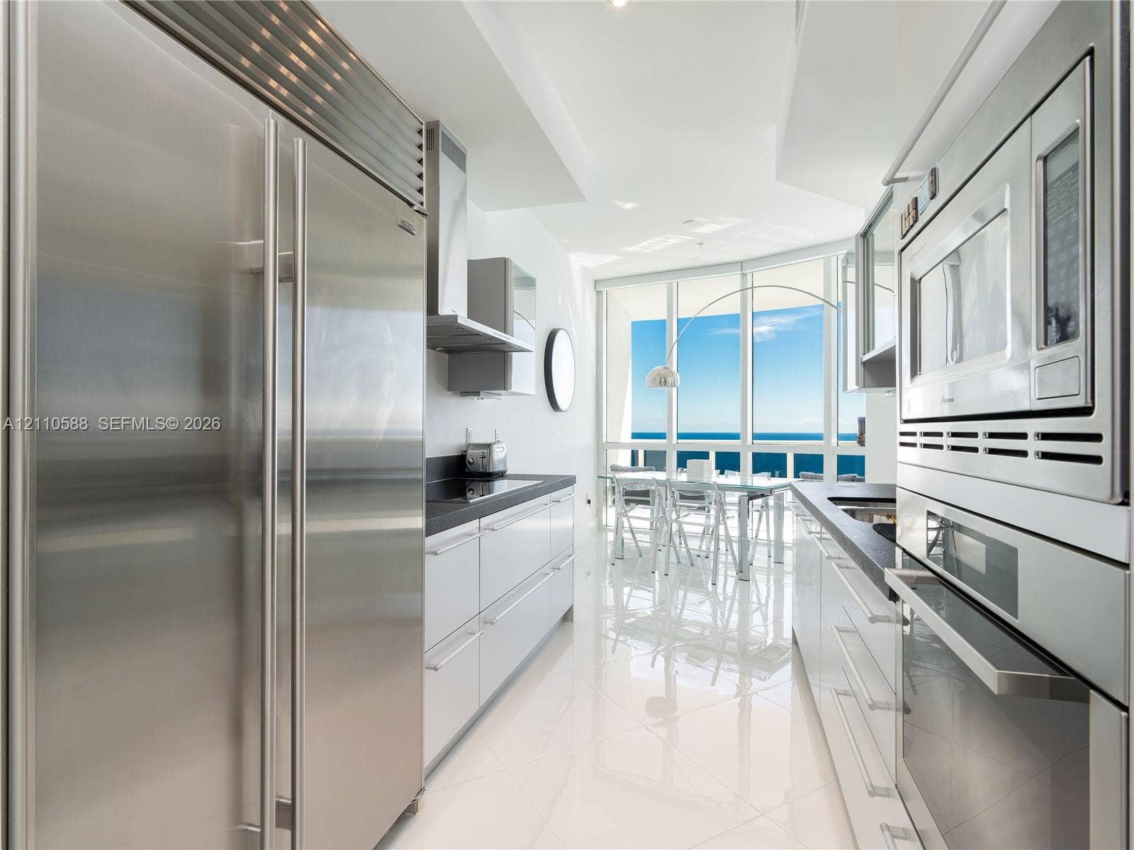 Modern galley kitchen featuring high-gloss cabinetry, stainless steel professional-grade appliances, dark stone countertops, polished tile flooring, and floor-to-ceiling windows