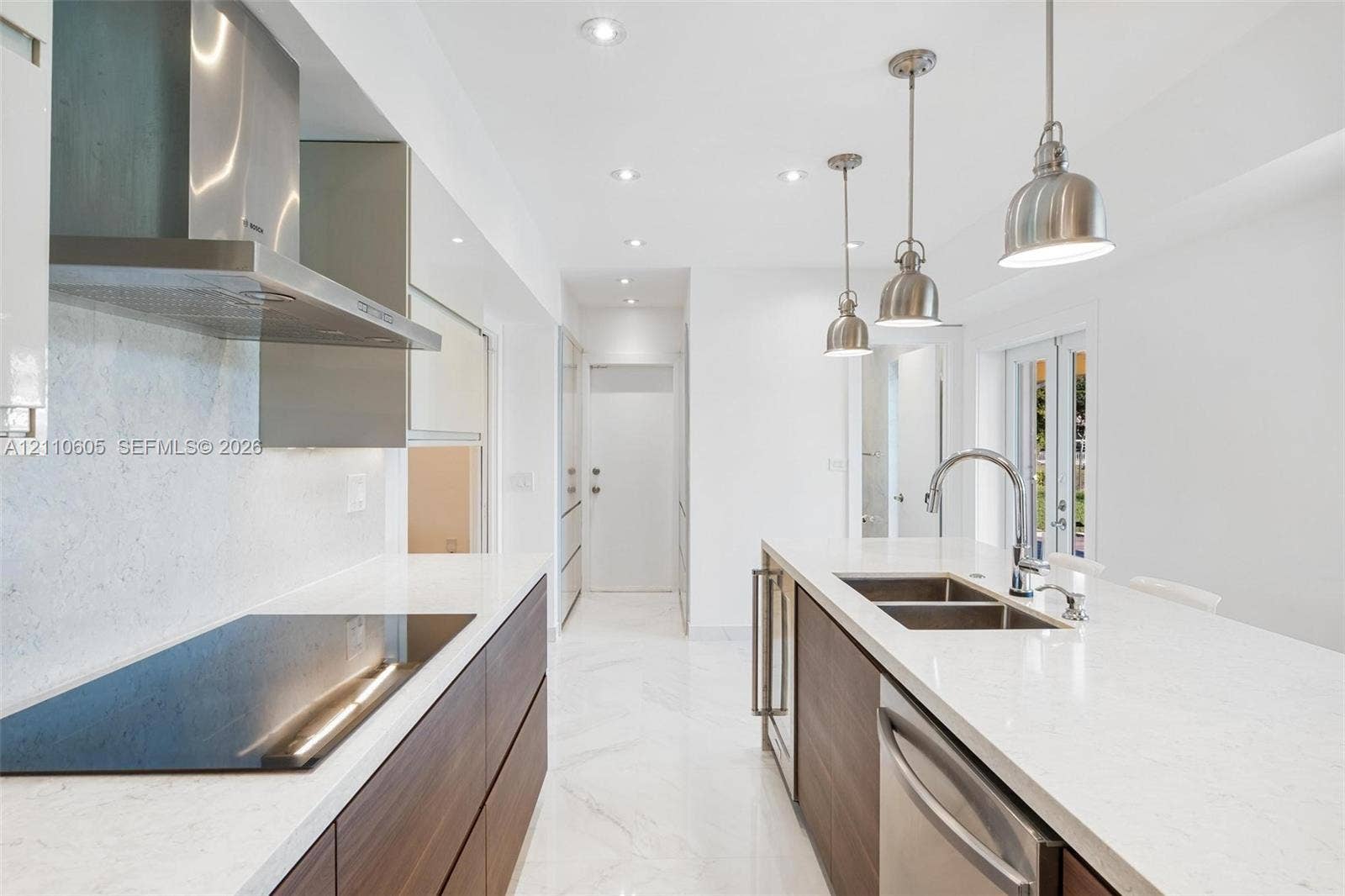 Modern kitchen featuring wood-finish flat-panel cabinetry, light-toned stone-look countertops, stainless steel appliances, marble-patterned flooring, and three pendant lights