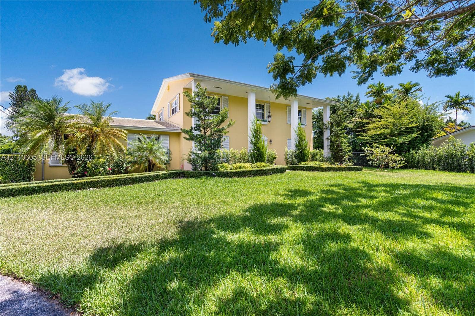 Two-story exterior featuring a columned front portico, light-toned stucco siding, and meticulously manicured hedging on an expansive lawn with mature palm trees