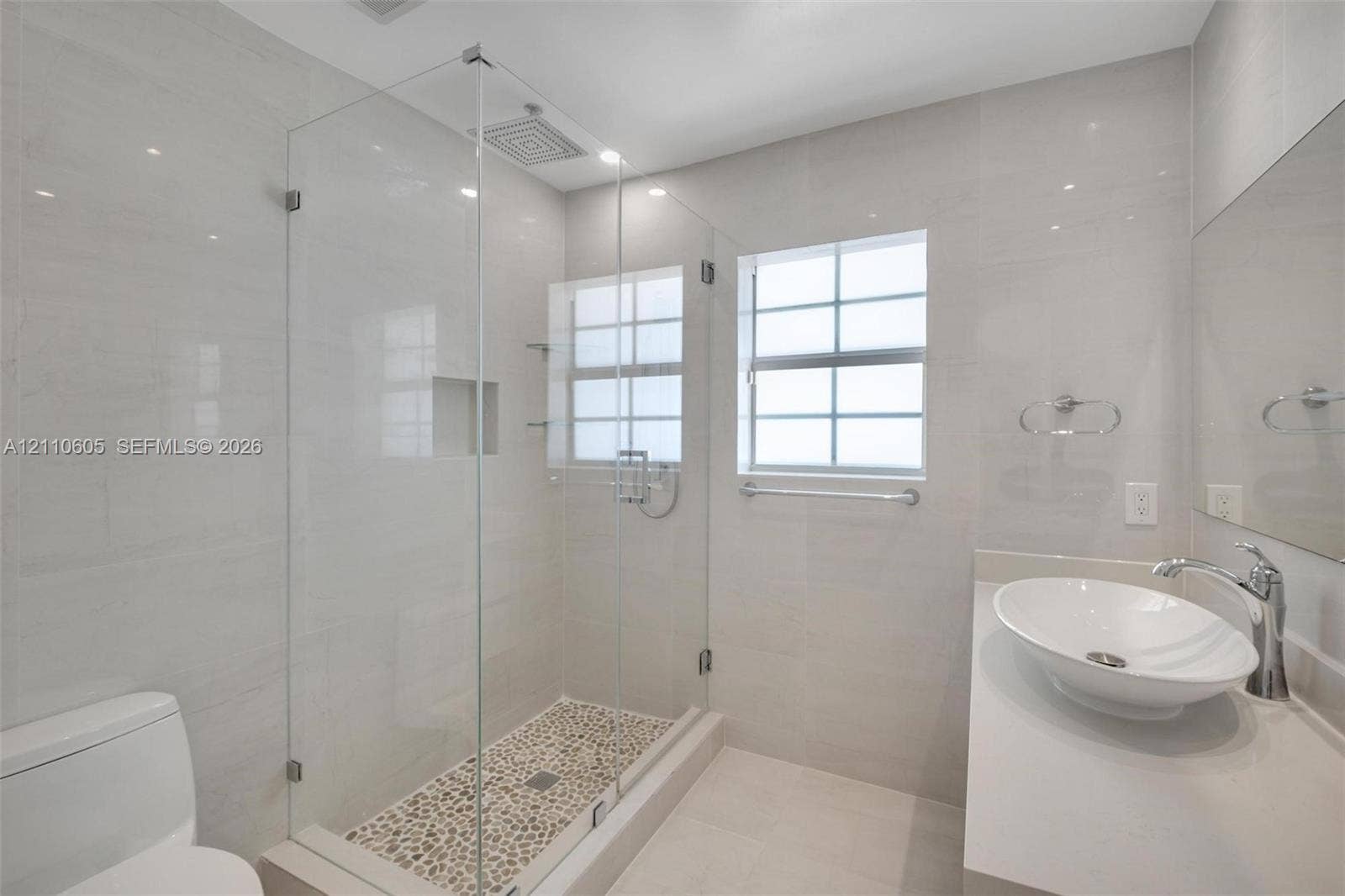 Bathroom featuring a glass-enclosed walk-in shower with pebble-stone flooring, a wall-mounted rainfall showerhead, a quartz-topped vanity with an oval vessel sink, and large-format porcelain wall tile