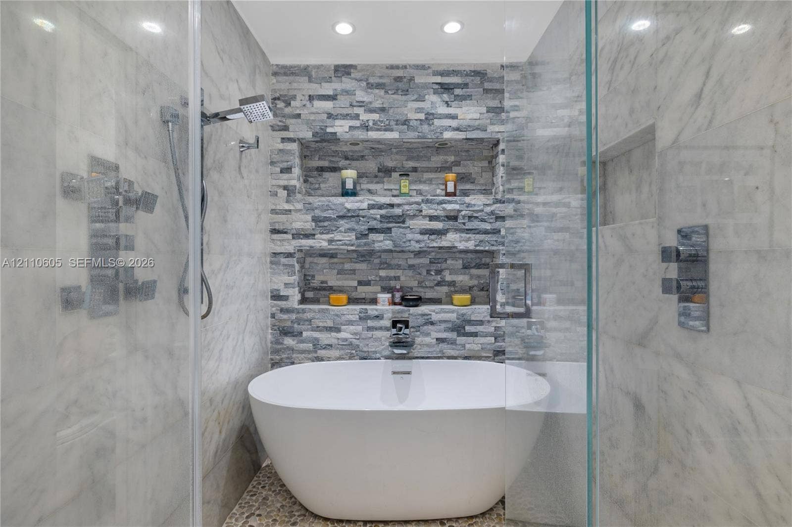 Freestanding soaking tub with polished chrome fixtures, integrated stacked-stone feature wall with dual recessed shelving, stone pebble flooring, and frameless glass shower enclosure