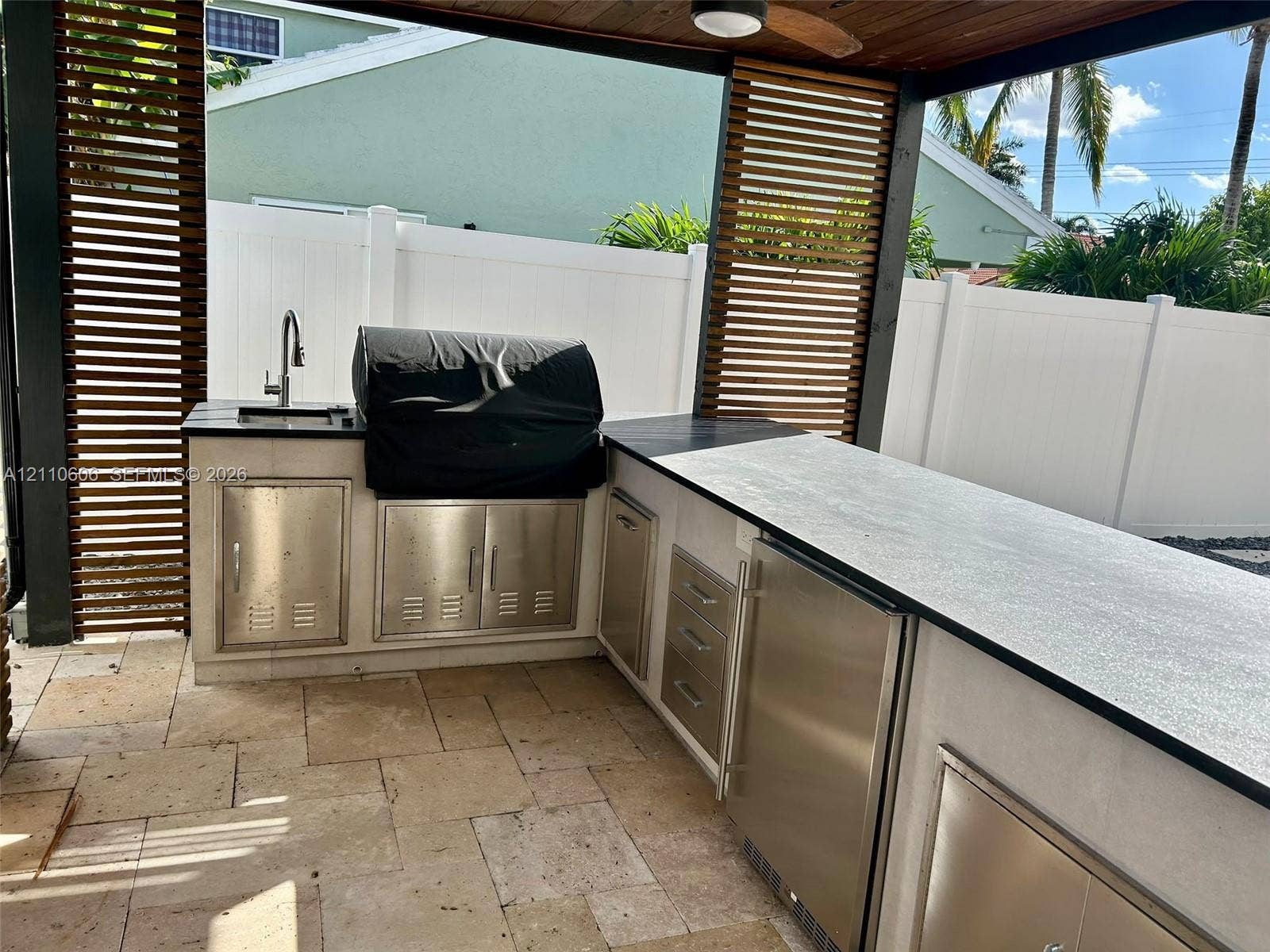 Covered outdoor kitchen featuring a stone-tile patio, built-in stainless steel cabinetry, a sink with a gooseneck faucet, and horizontal wood-slat privacy screens