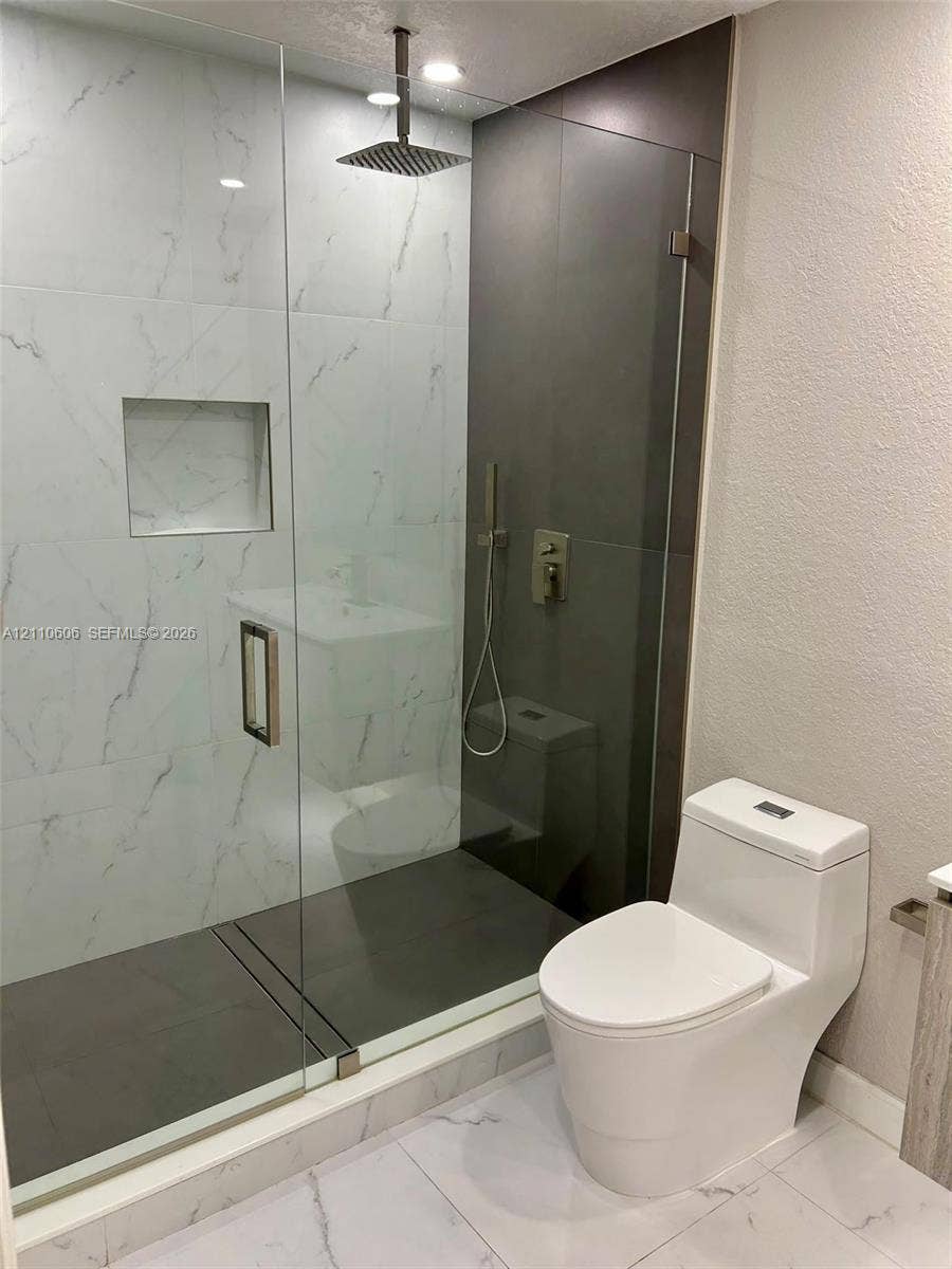Walk-in shower featuring a frameless glass enclosure, rainfall showerhead, recessed storage niche, and contrast stone-finish tiling, paired with a modern dual-flush elongated toilet